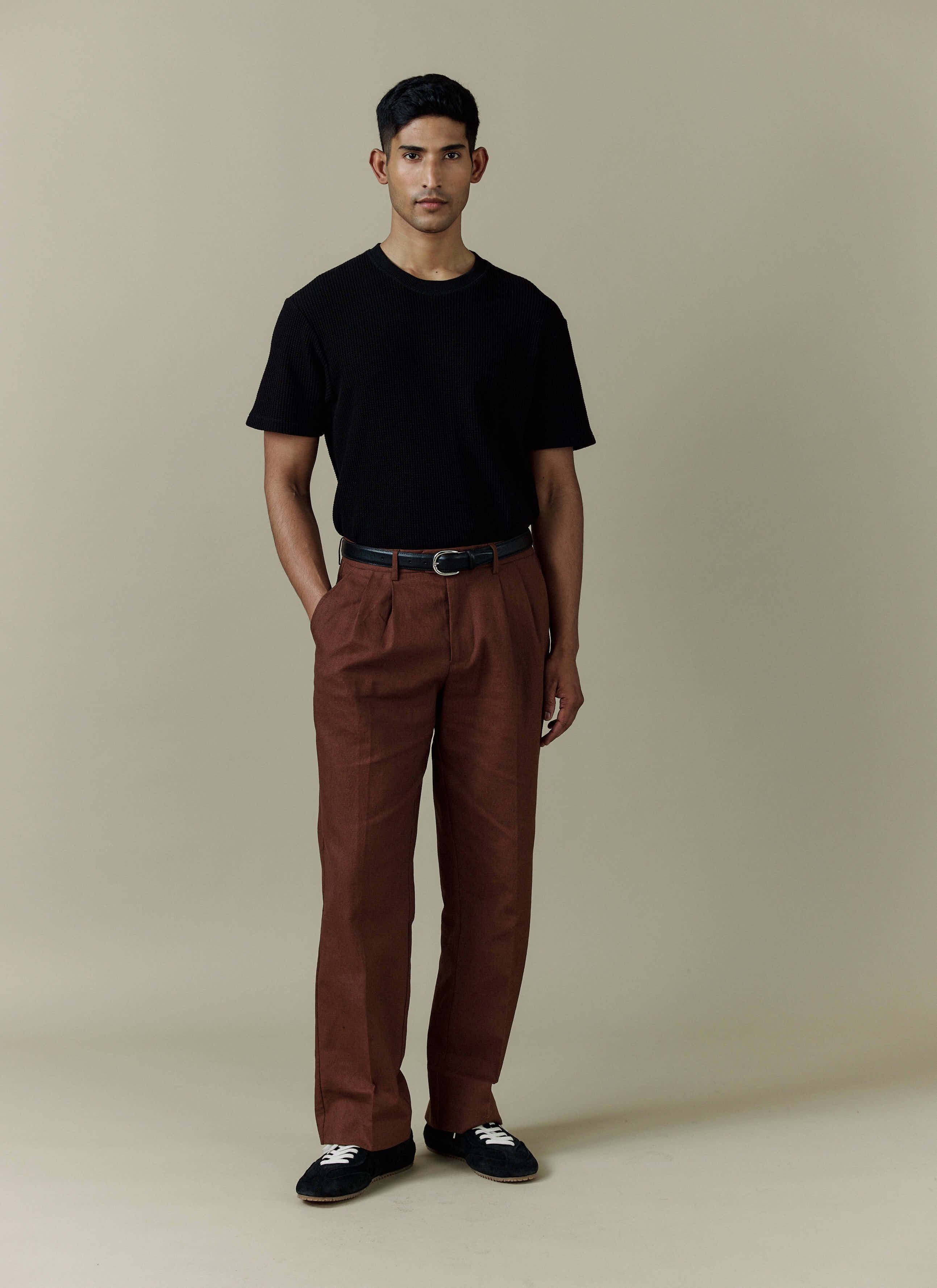A model wears the brown linen Pleated Tailored Trousers from Percival with a black t-shirt and trainers.