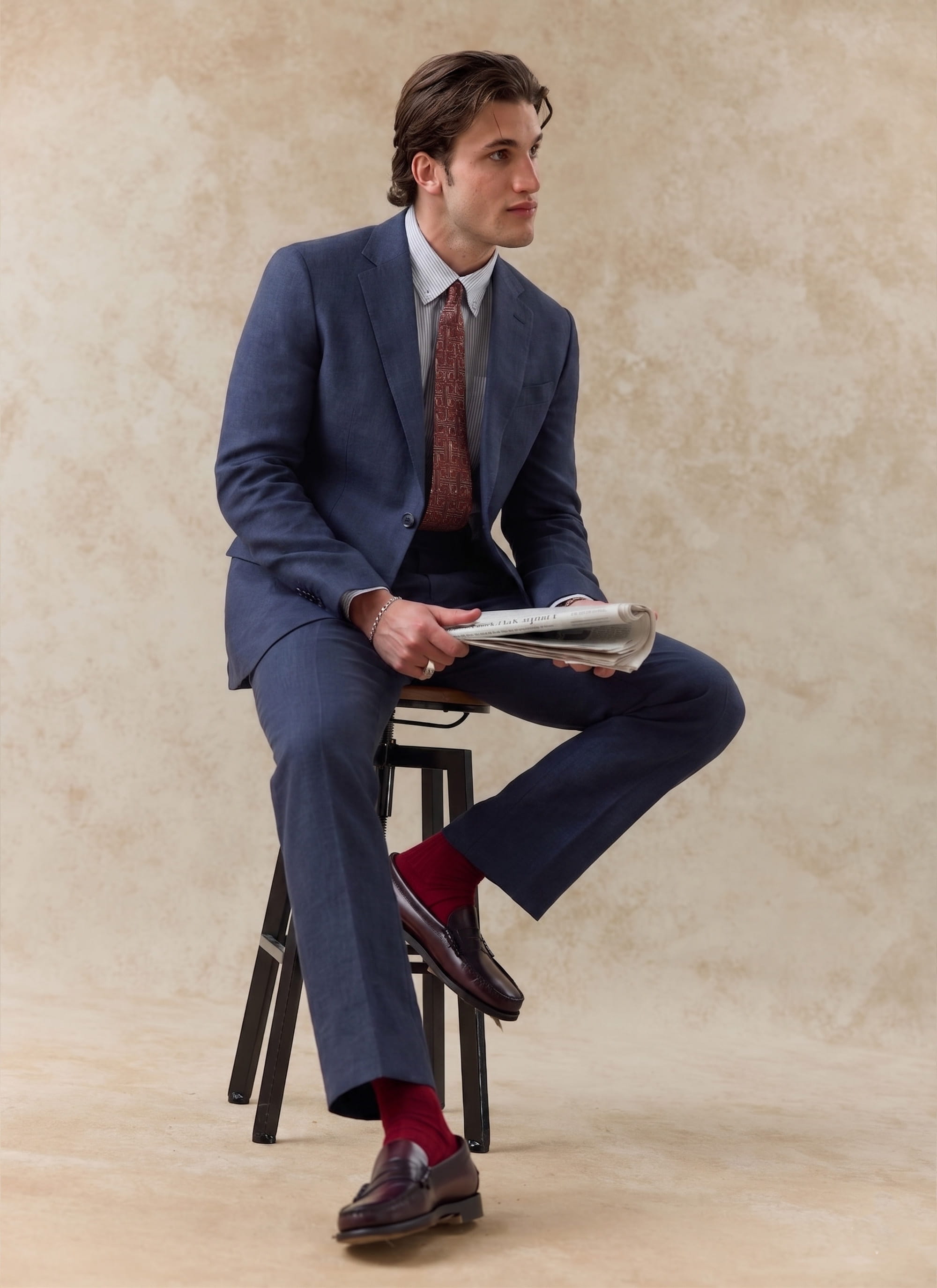 Navy blue Percival Tailored Linen Blazer and matching suit trousers worn by a seated model holding a newspaper.