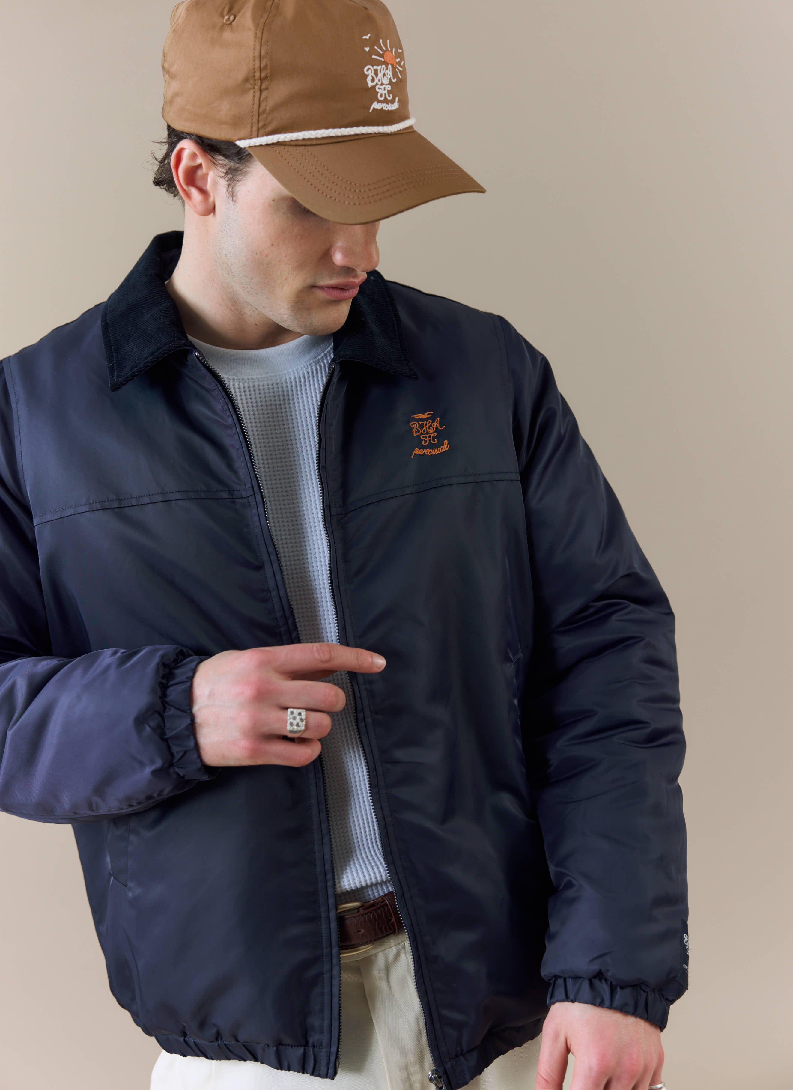 A model wears the Percival x Brighton & Hove Albion F.C. Coach Jacket in navy, showing the orange chest embroidery.