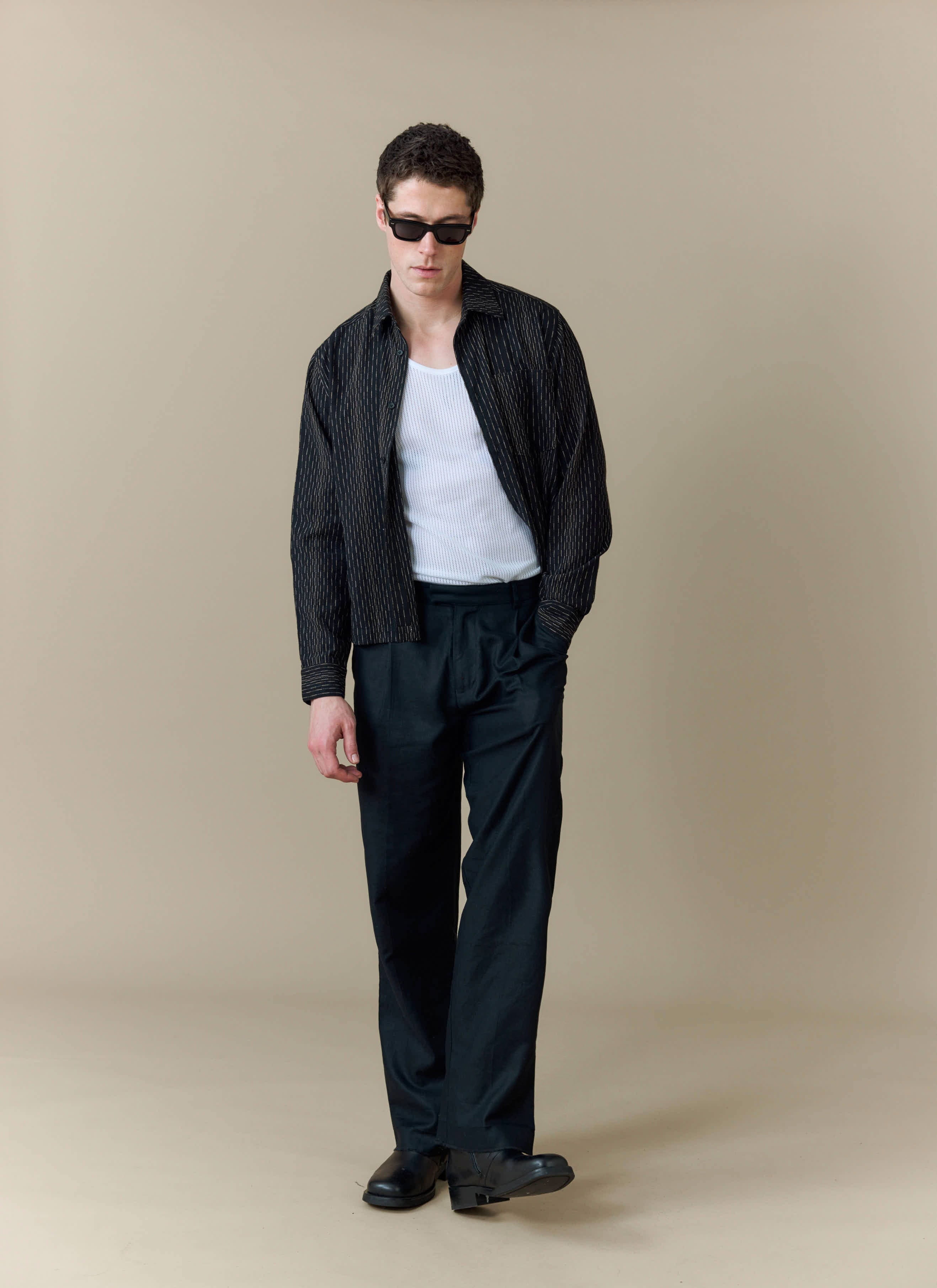 A model wears the Percival Leland Cuban Boxy Shirt in black, styled open over a white vest with black trousers & sunglasses.