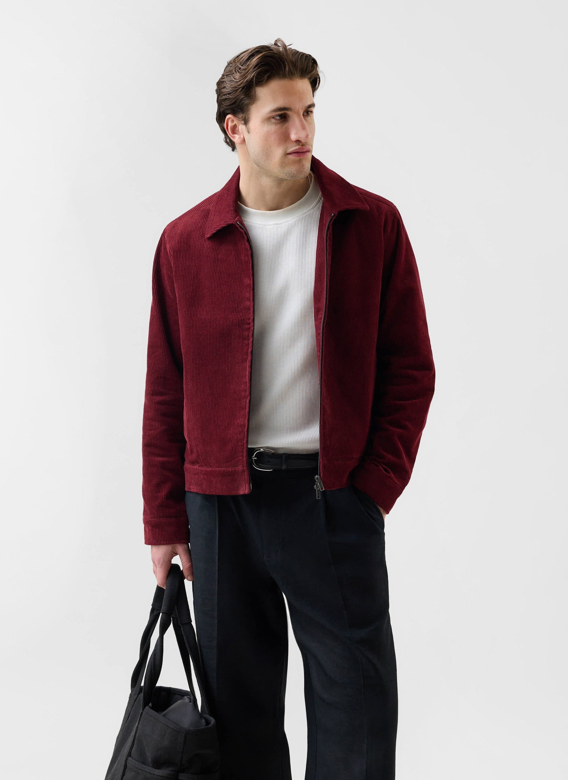 A model wears the Percival Marcel Cord Jacket in burgundy, styled with a white t-shirt, dark trousers, and a black bag.