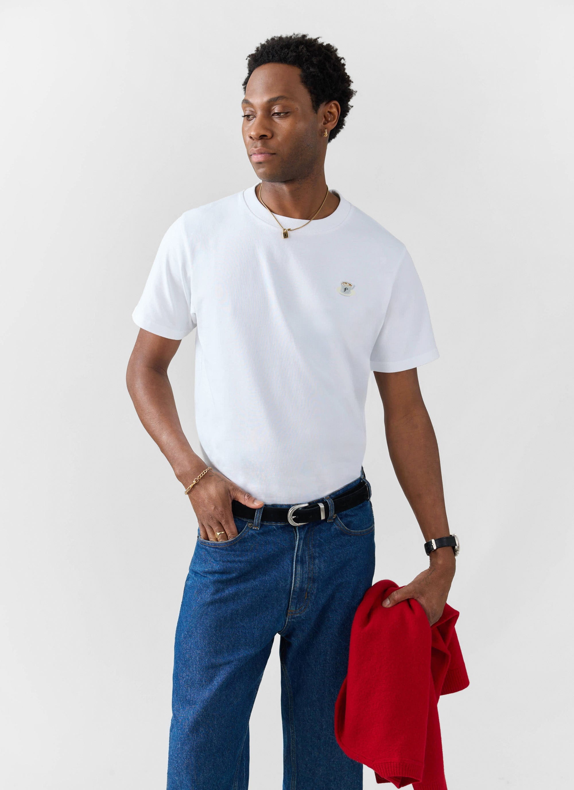 A model wearing the white Percival Latte Embroidered T Shirt, paired with blue jeans and holding a red jumper.