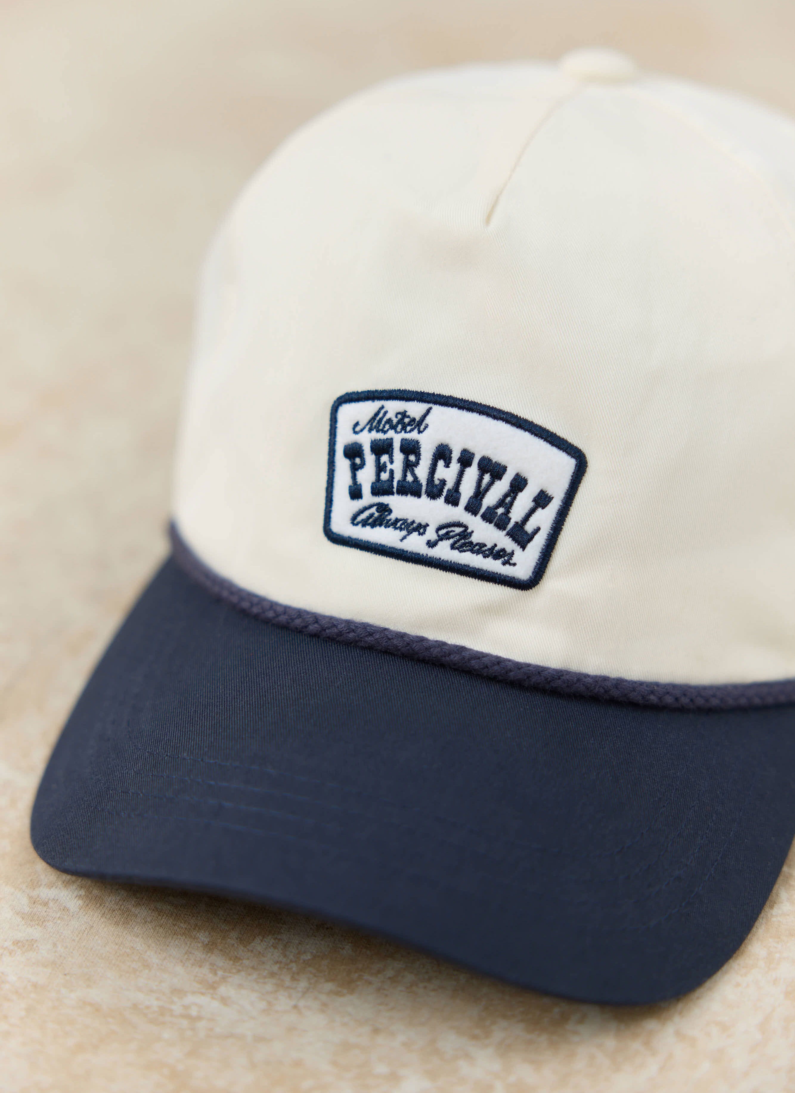 Close-up of the cream Percival Donovan soft trucker cap, featuring a navy brim and an embroidered motel-inspired patch.