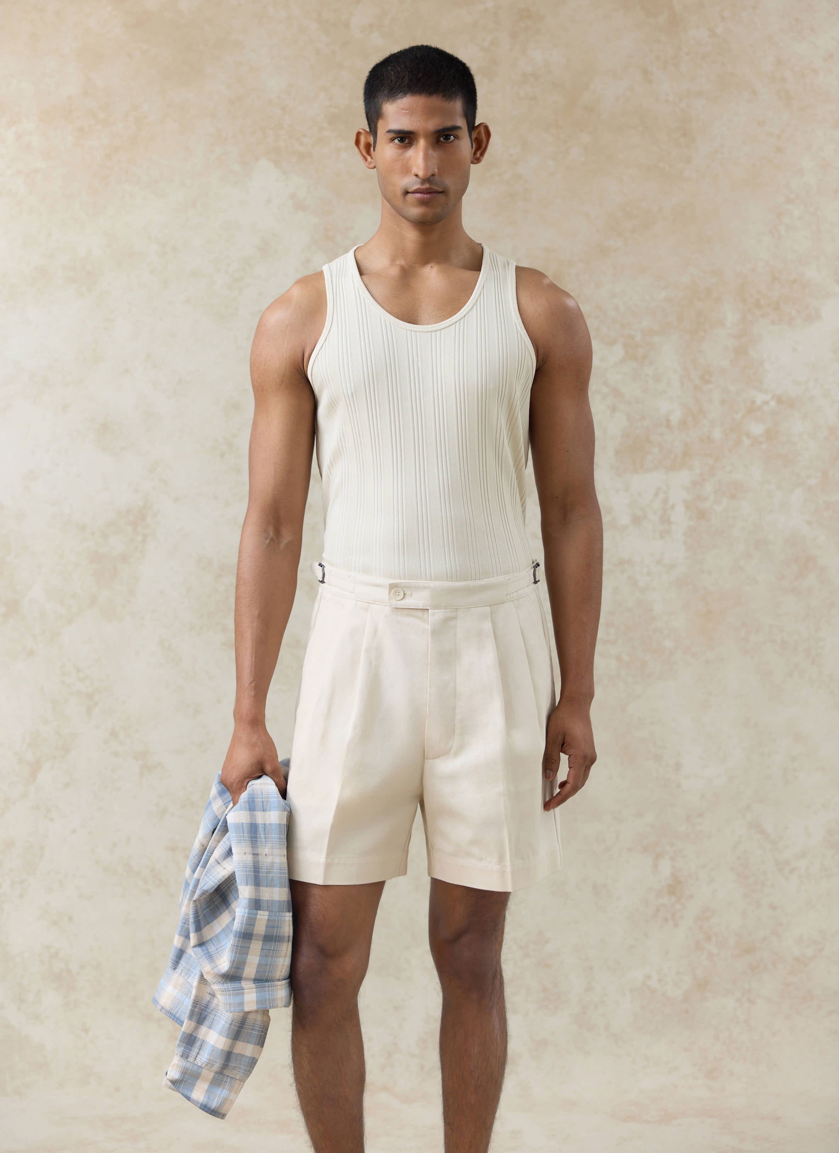 Model wears the cream Percival Leon Ribbed Vest and matching Blake double pleated cotton shorts, holding a checked shirt.