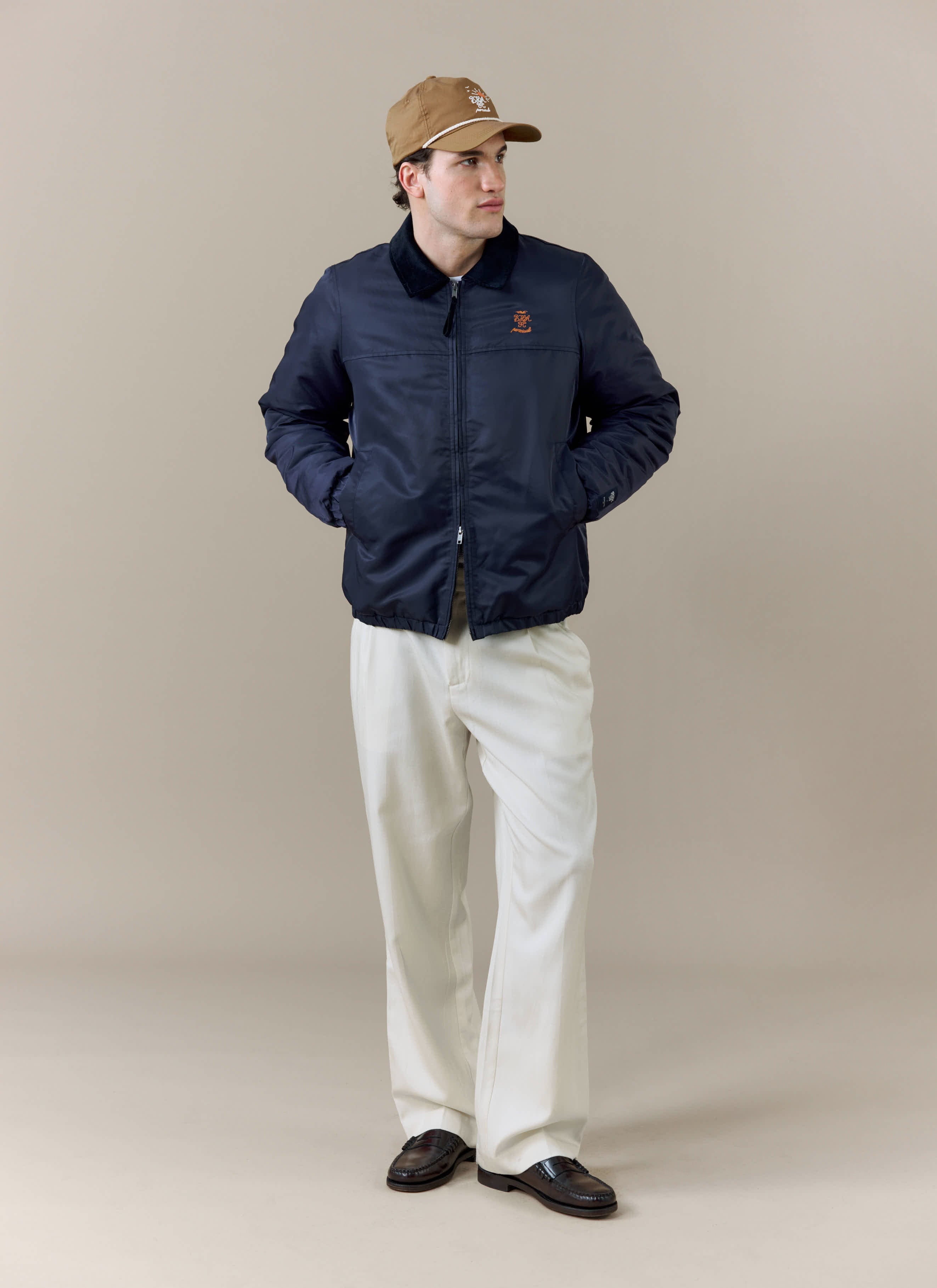 A model wears the Percival x Brighton & Hove Albion F.C. navy Coach Jacket, styled with white trousers and a tan cap.