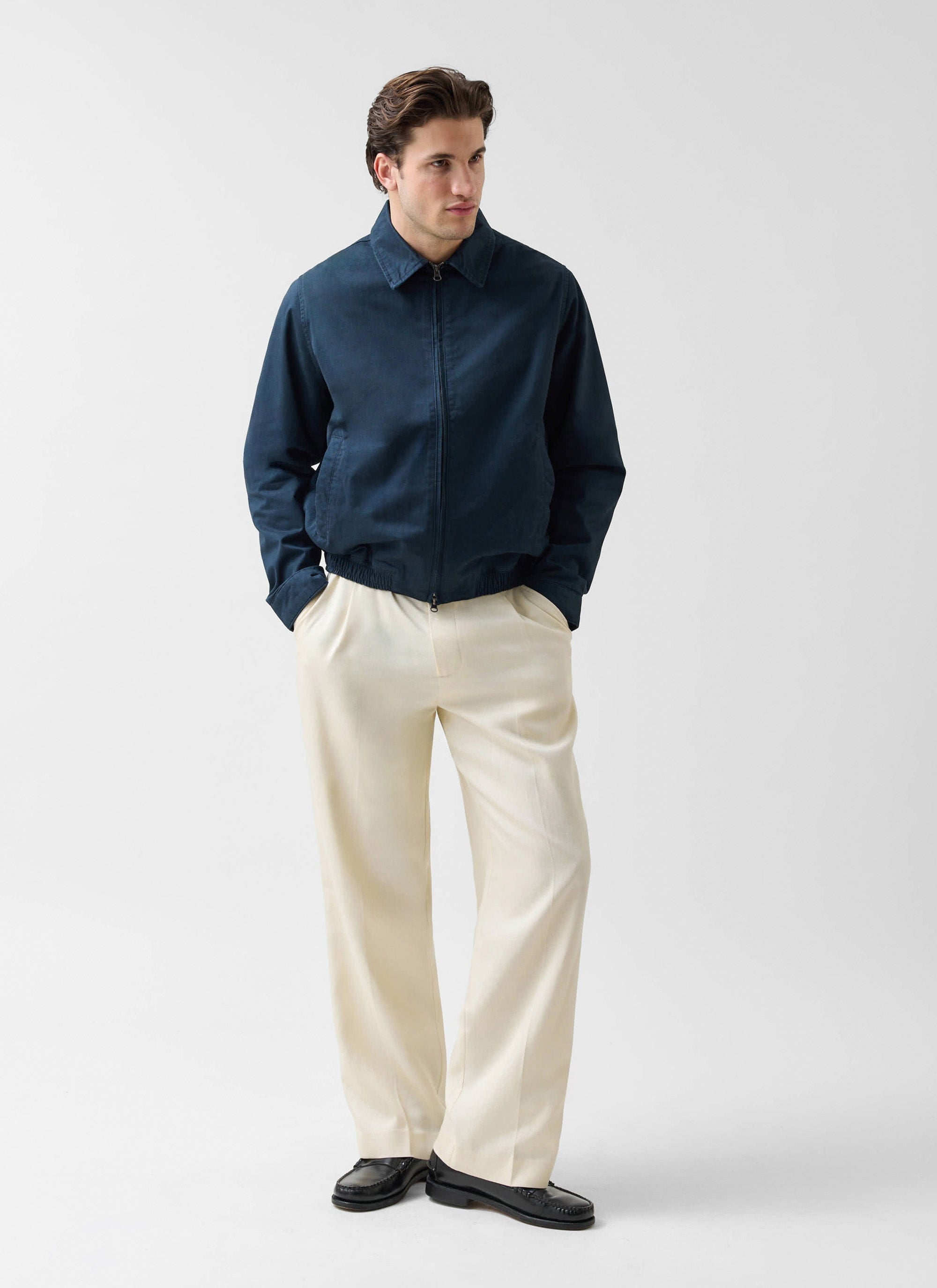 A model wears the Percival Leo Harrington Jacket in navy cotton, styled with cream trousers and black loafers.