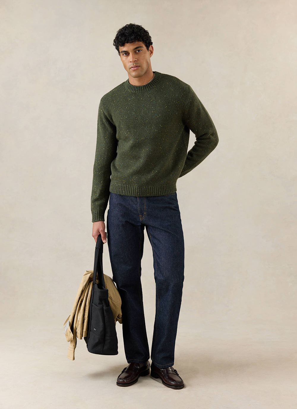 A model wears the forest green Percival Rhodes crew neck jumper with dark denim jeans and loafers, holding a bag and jacket.