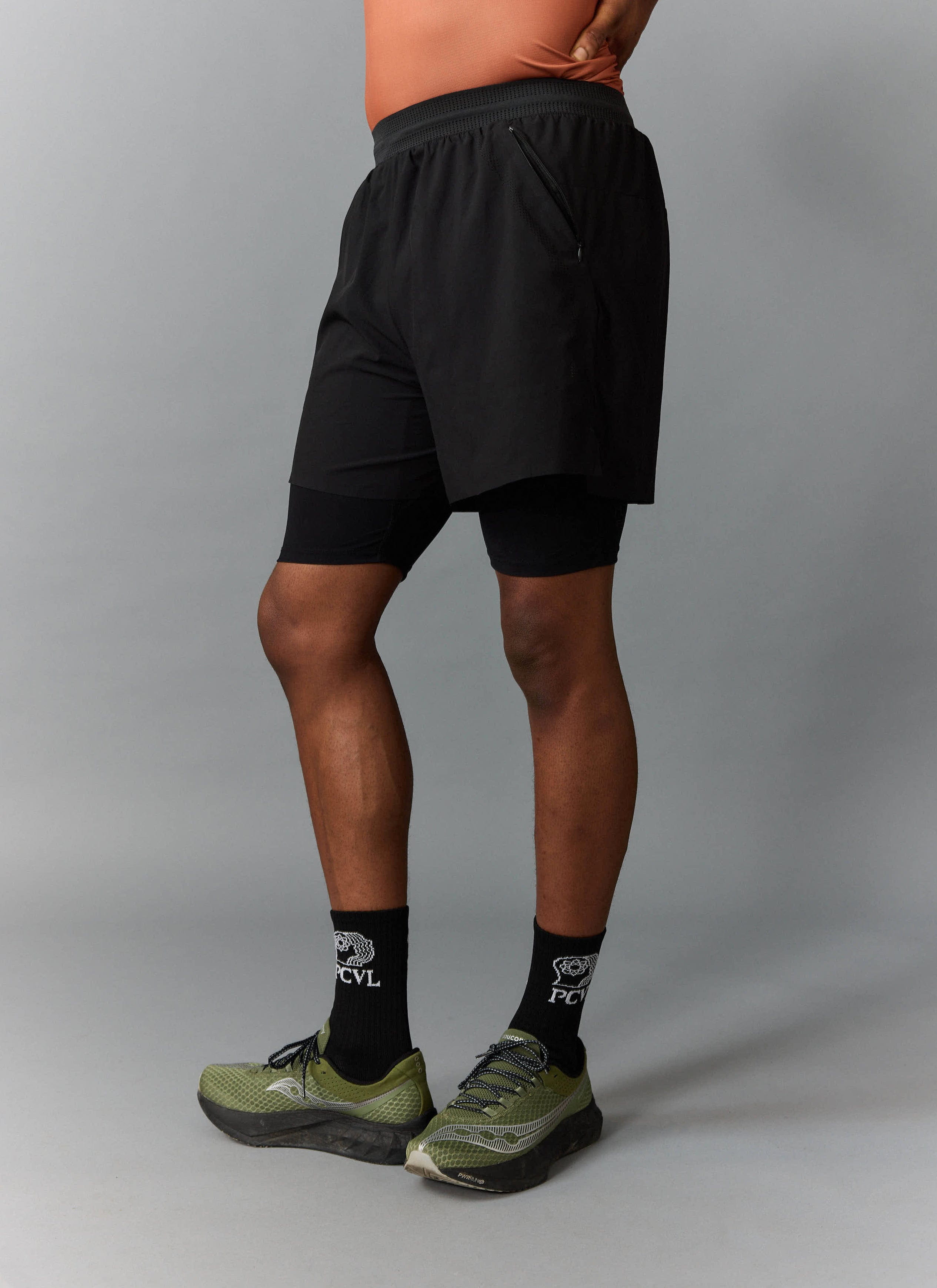 A side view of a model wearing the black PCVL 2 in 1 performance shorts from Percival with matching socks.