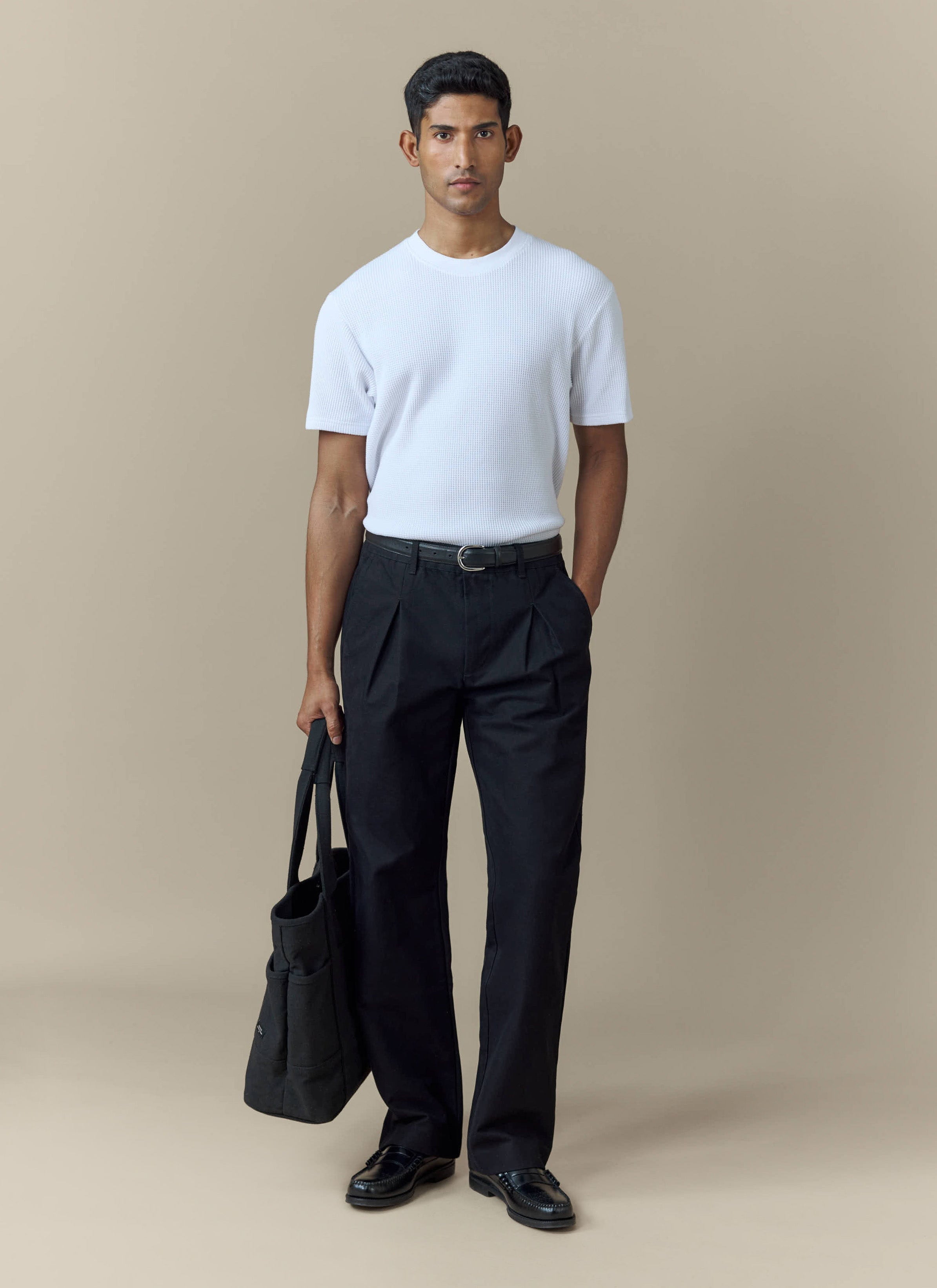 A model wears the Percival Seb Inverted Pleat Trousers in black, styled with a white ribbed t-shirt and black loafers.
