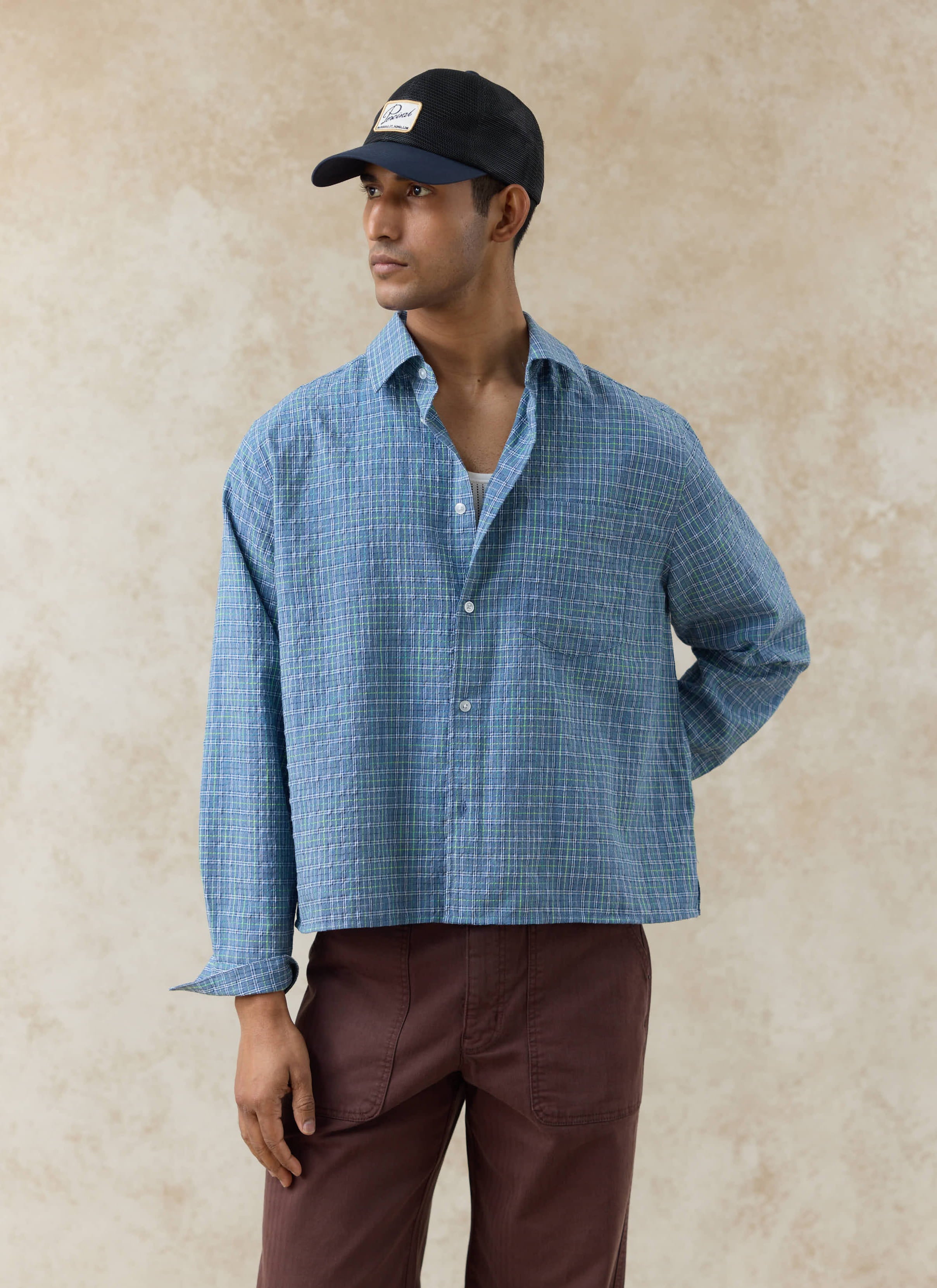 Percival Hinsley Checked Boxy Shirt in blue cotton grid, styled on a model with brown trousers and a black mesh cap.