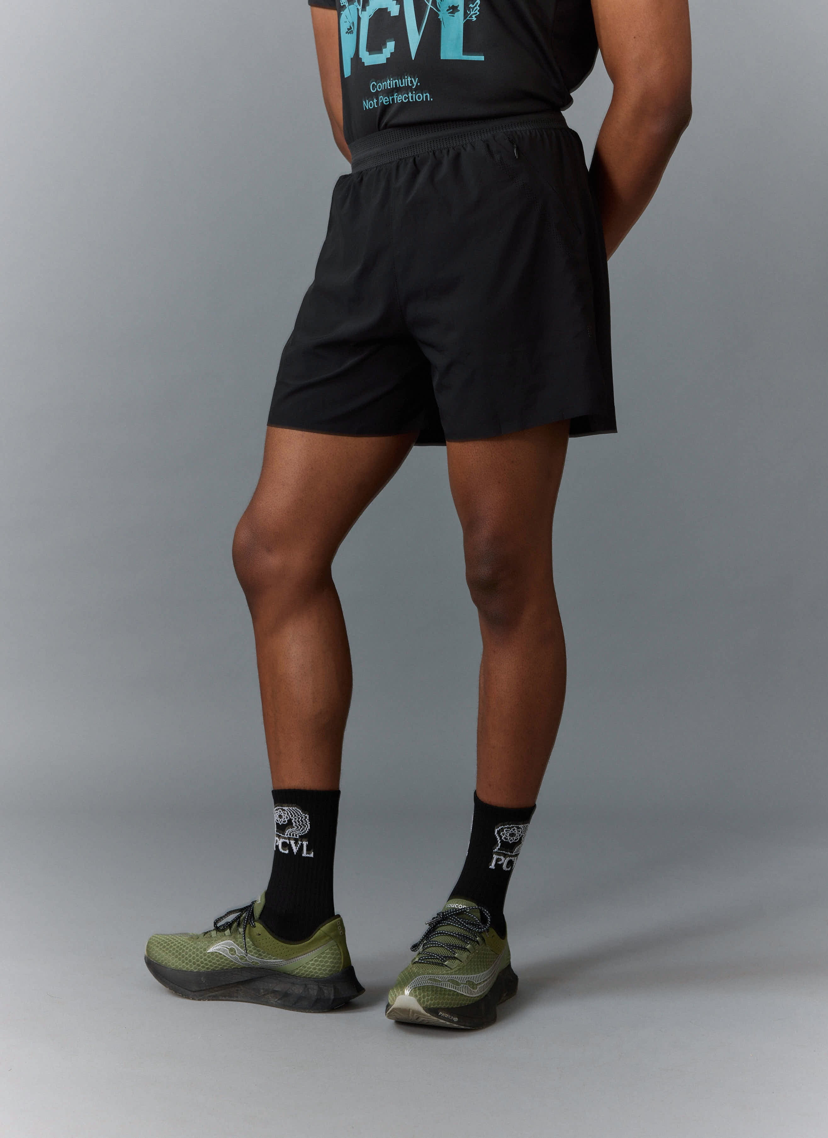 A model wears the black PCVL Performance Shorts from Percival, seen from the side with matching PCVL socks and green shoes.