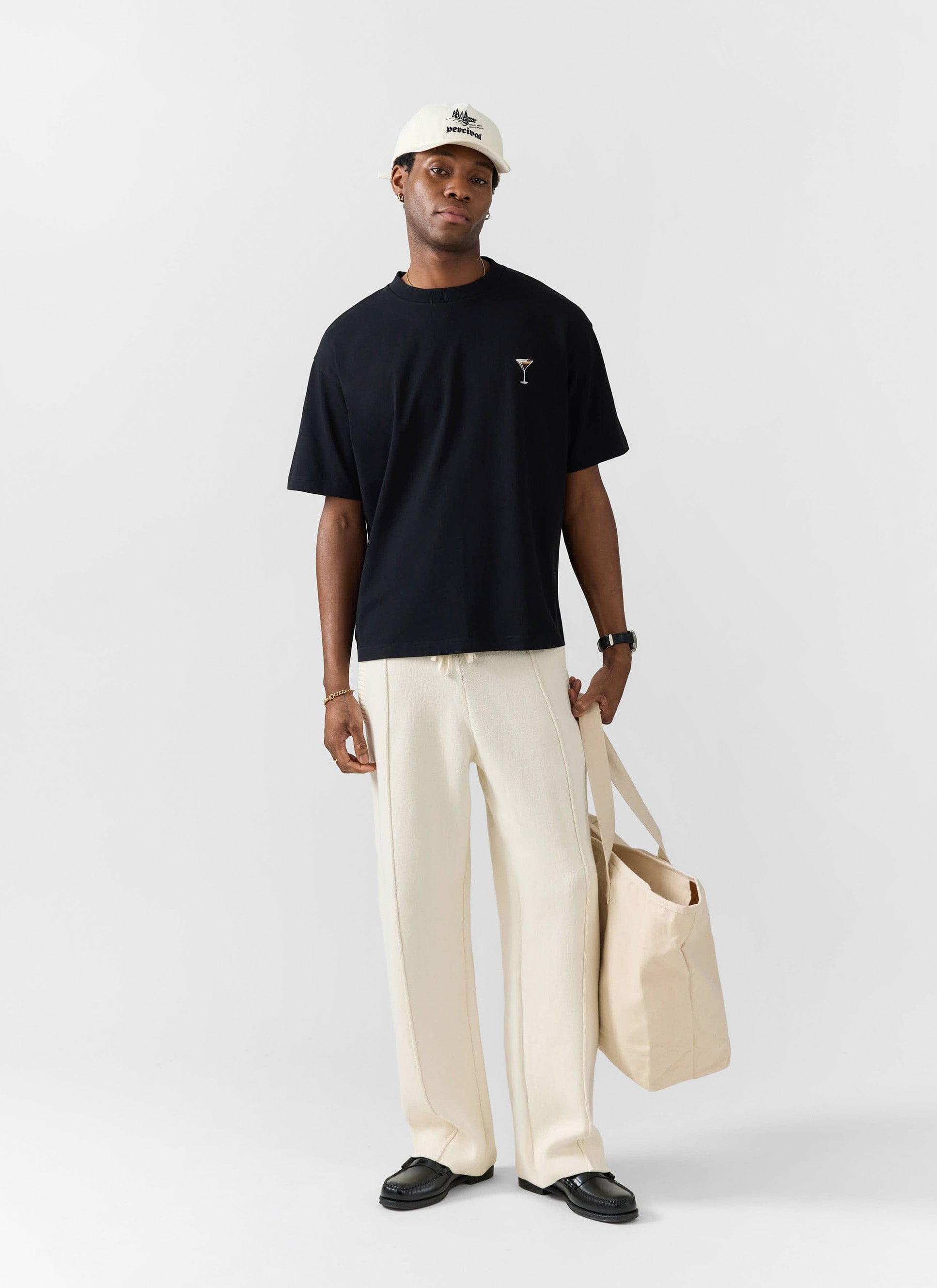 A model wears the black Percival Espresso Martini embroidered T-shirt, styled with cream trousers and holding a tote bag.