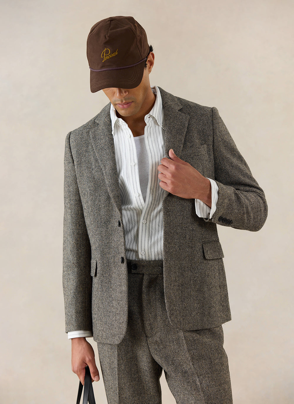 A model wears the Percival Wilde Tailored Blazer in Birdseye Wool Grey, styled with a striped shirt and a brown cap.