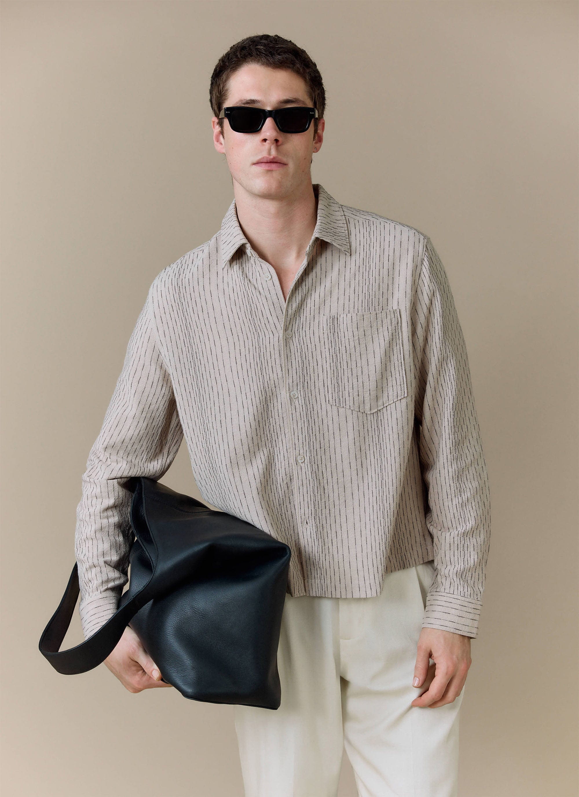 A model wears the ecru Leland Cuban Boxy Shirt from Percival with white trousers, sunglasses and a black bag.