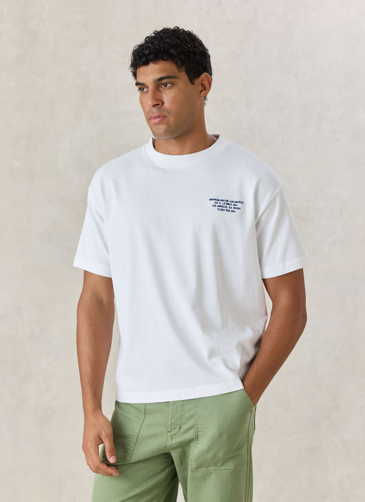 Model wearing the white American Rag x Percival oversized T-shirt featuring navy chest lettering, paired with green trousers.