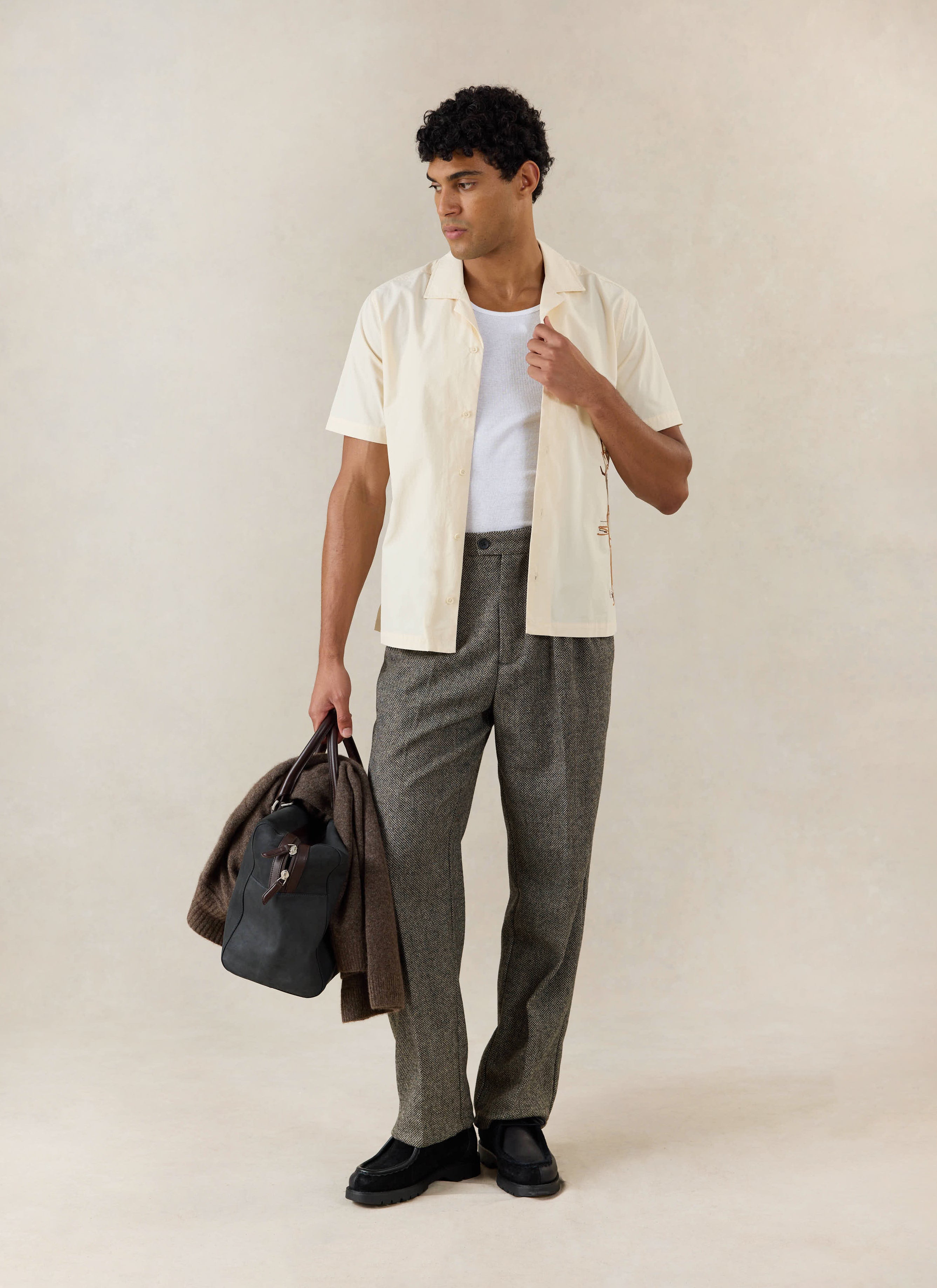 A model wears the cream Percival Supper Club Embroidered Cuban Shirt over a white vest, with grey trousers and a holdall.