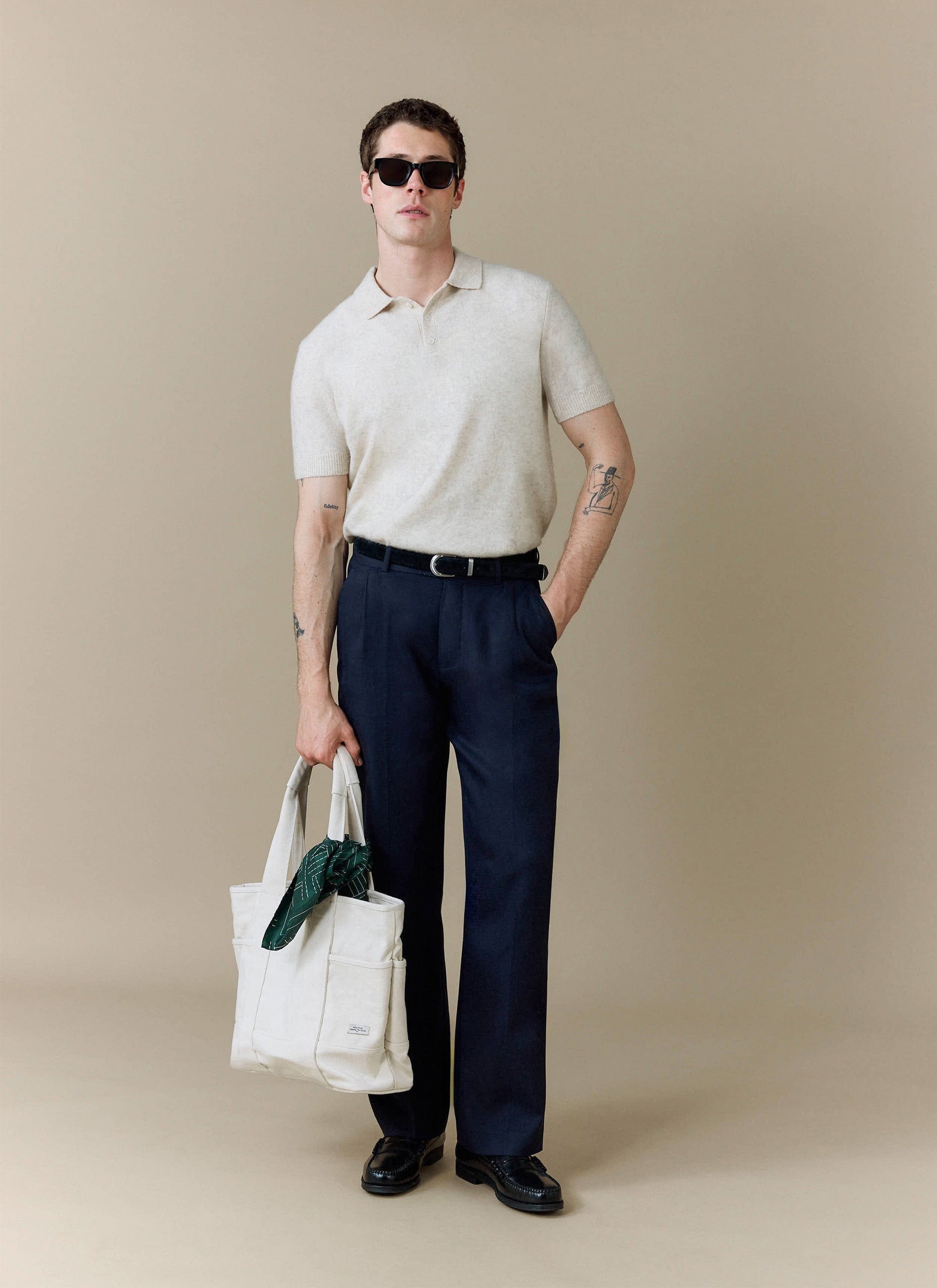 A model wears the Percival Collins Boxy Knitted Polo in ecru, styled with dark trousers, sunglasses, and a tote bag.