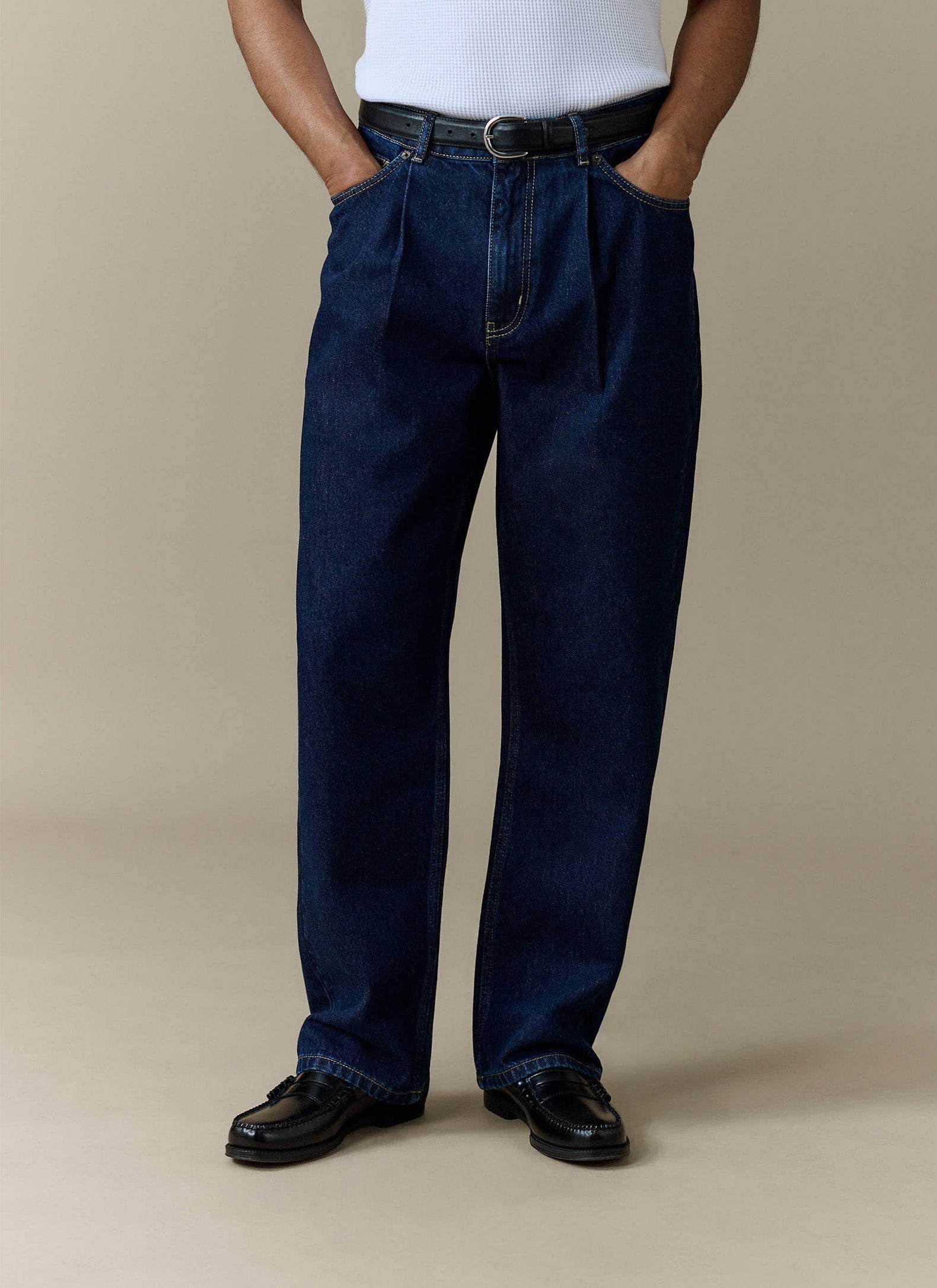 A model wears the Percival Pleated Denim Jeans in dark indigo, styled with a white top and black loafers.