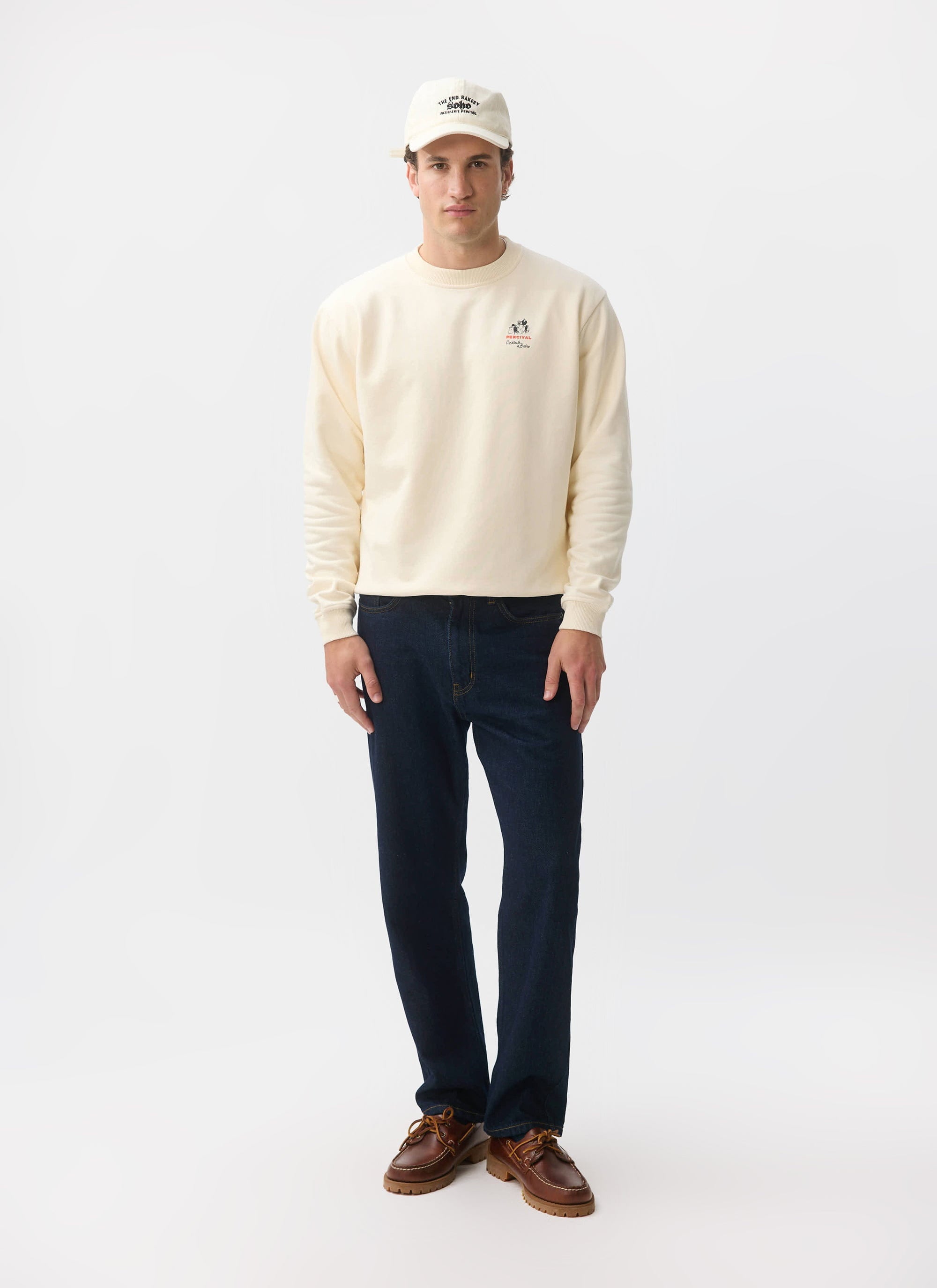 A model wears the Percival Bistro Printed Sweatshirt in ecru, styled with dark blue jeans, brown shoes and a matching cap.