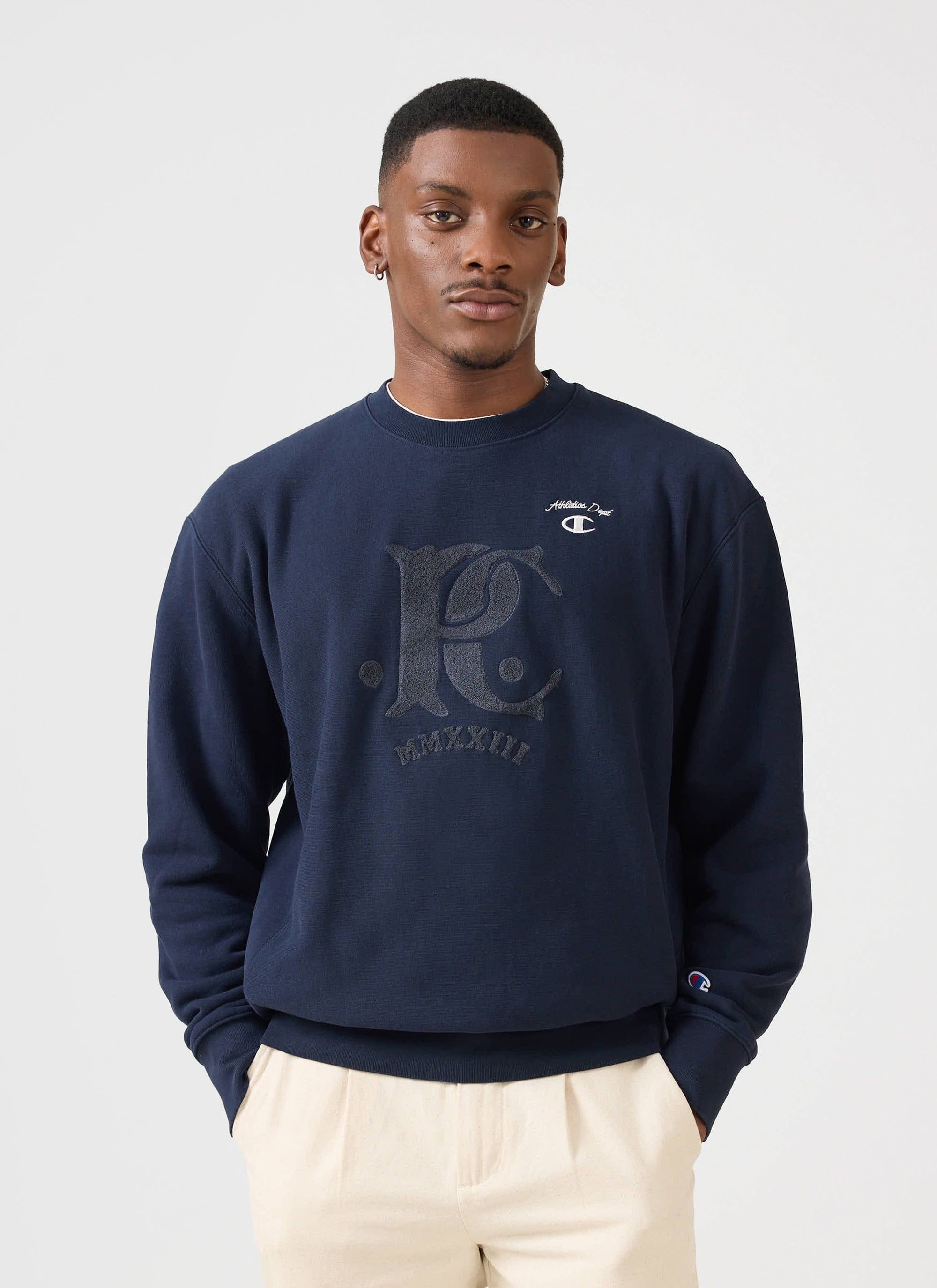 A model wears the navy Champion x Percival jersey sweatshirt, featuring a large tonal chenille patch.