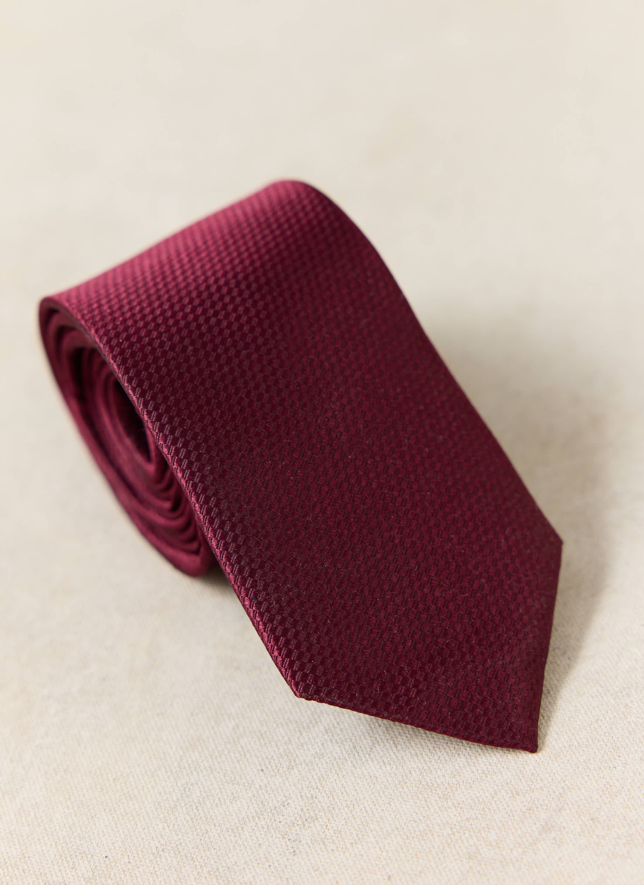 The Percival Grenadine Tie in Red Silk, featuring a textured weave, shown rolled up on a neutral background.