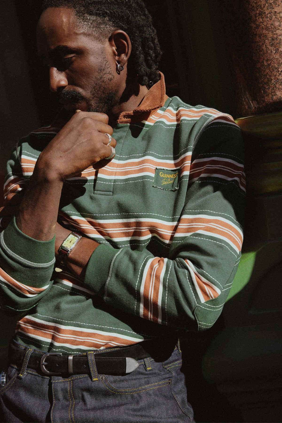 Model wearing the Guinness x Percival striped rugby polo shirt in forest green, showing cord collar and chest appliqué.