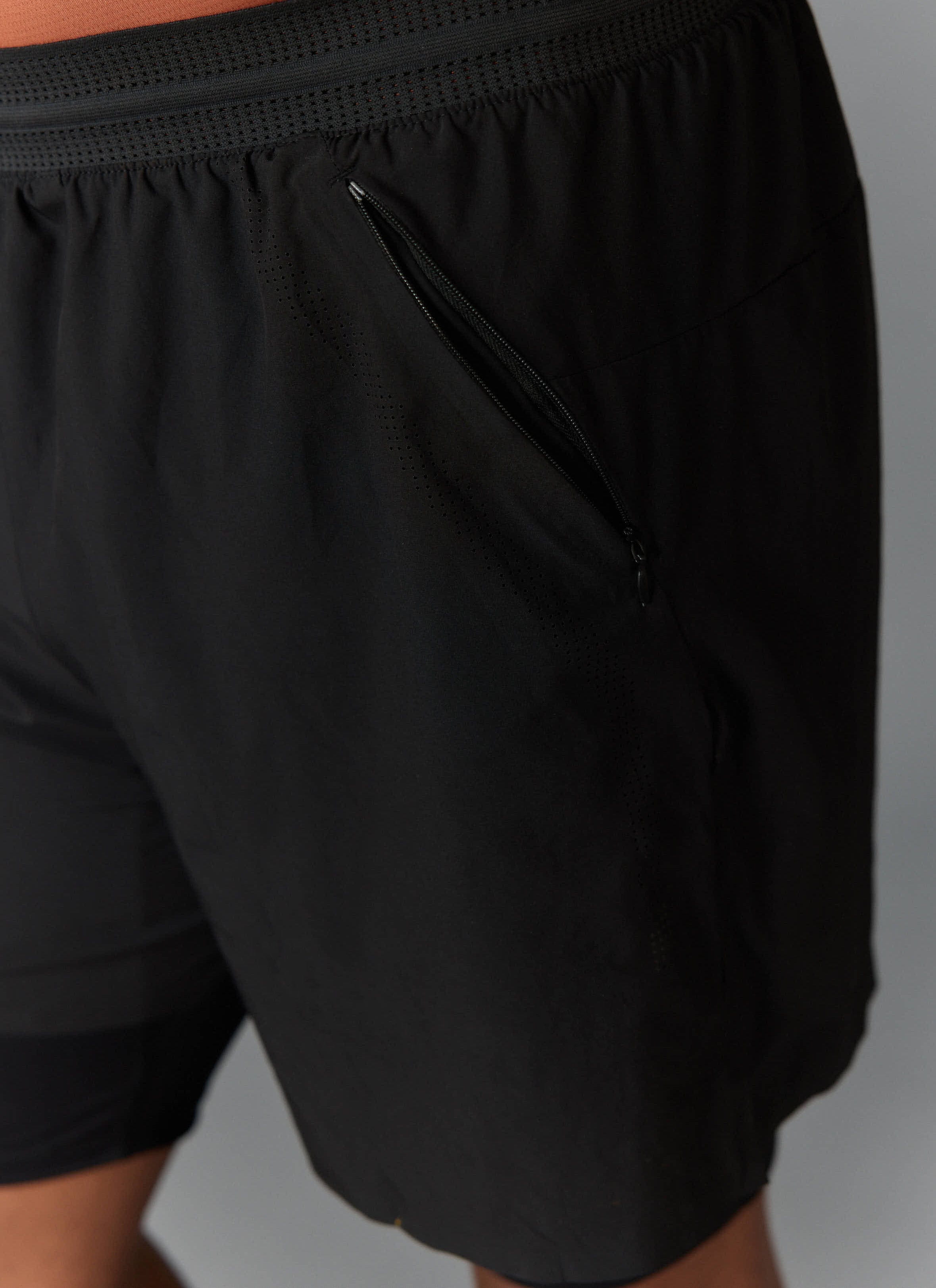 A close-up of the black PCVL 2 in 1 Shorts from Percival, showing the perforated waistband and secure zipped pocket detail.
