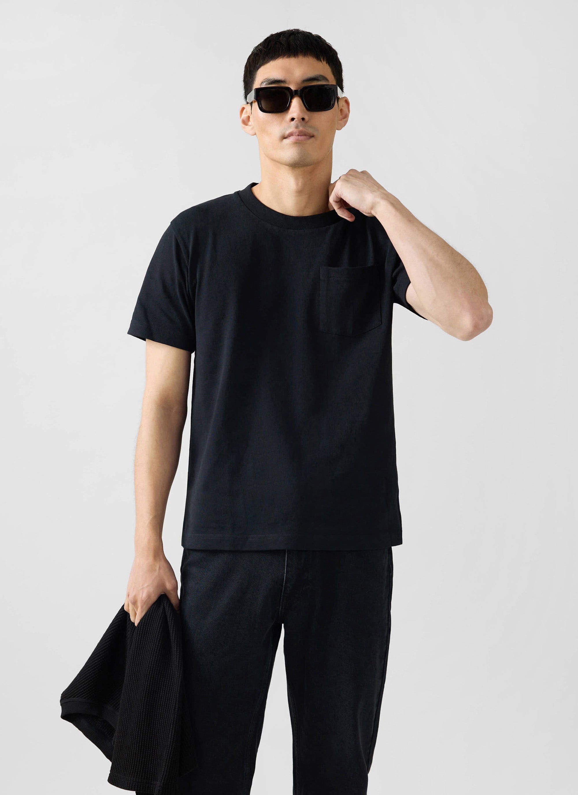 A man models the Percival black organic cotton Short Sleeve Pocket T-Shirt with sunglasses against a studio background.