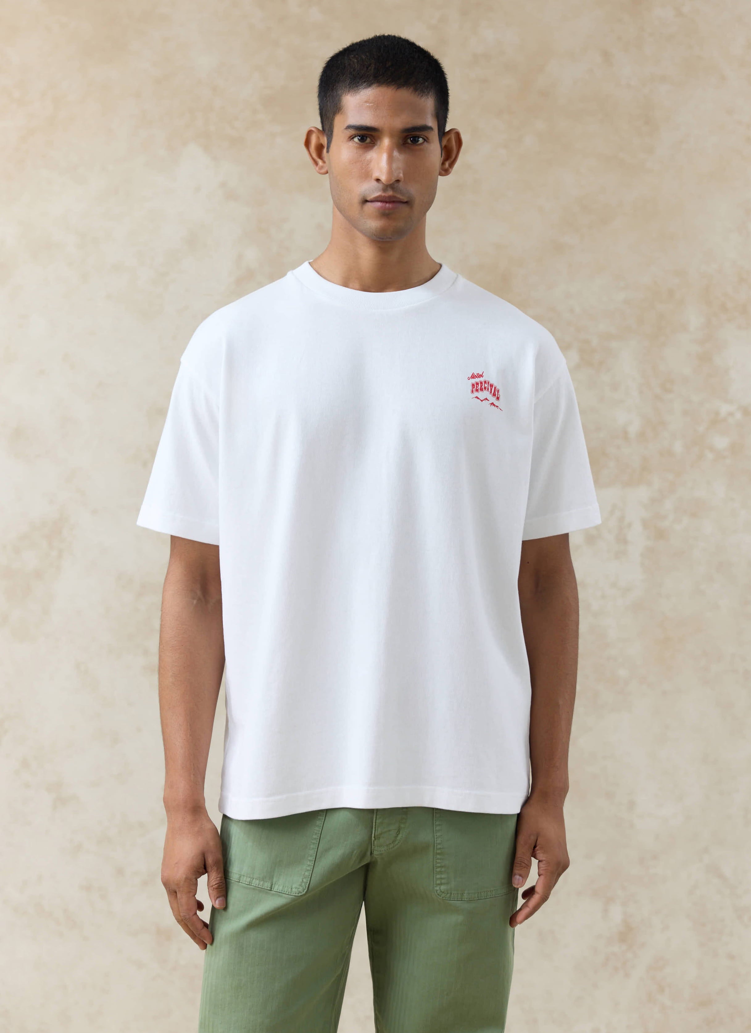 White organic cotton Gordon Oversized T Shirt by Percival, with red Motel Percival chest print, worn with green trousers.
