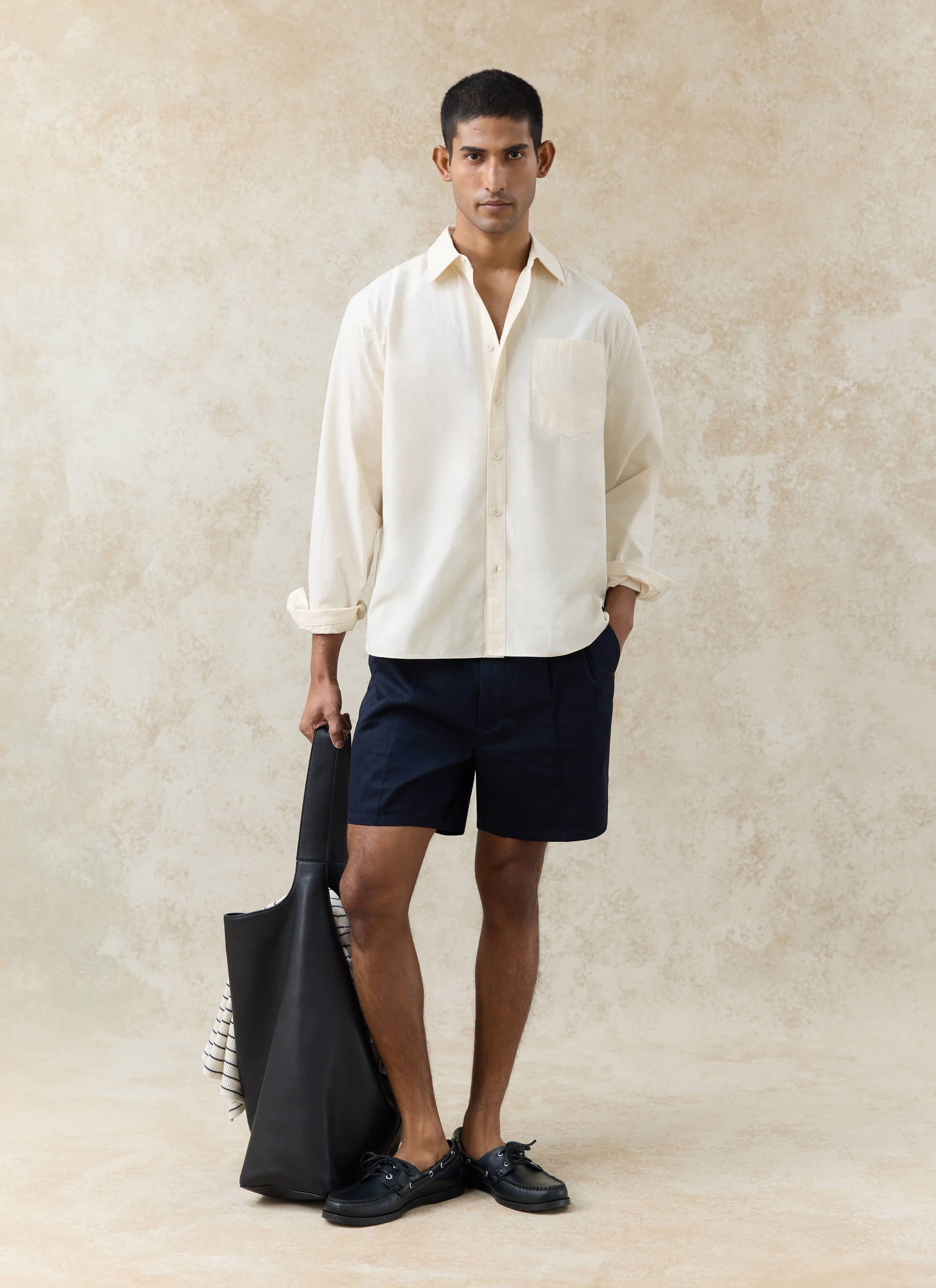Model wearing the Percival Dali Relaxed Poplin Shirt in cream cotton, paired with navy shorts and a black tote bag.