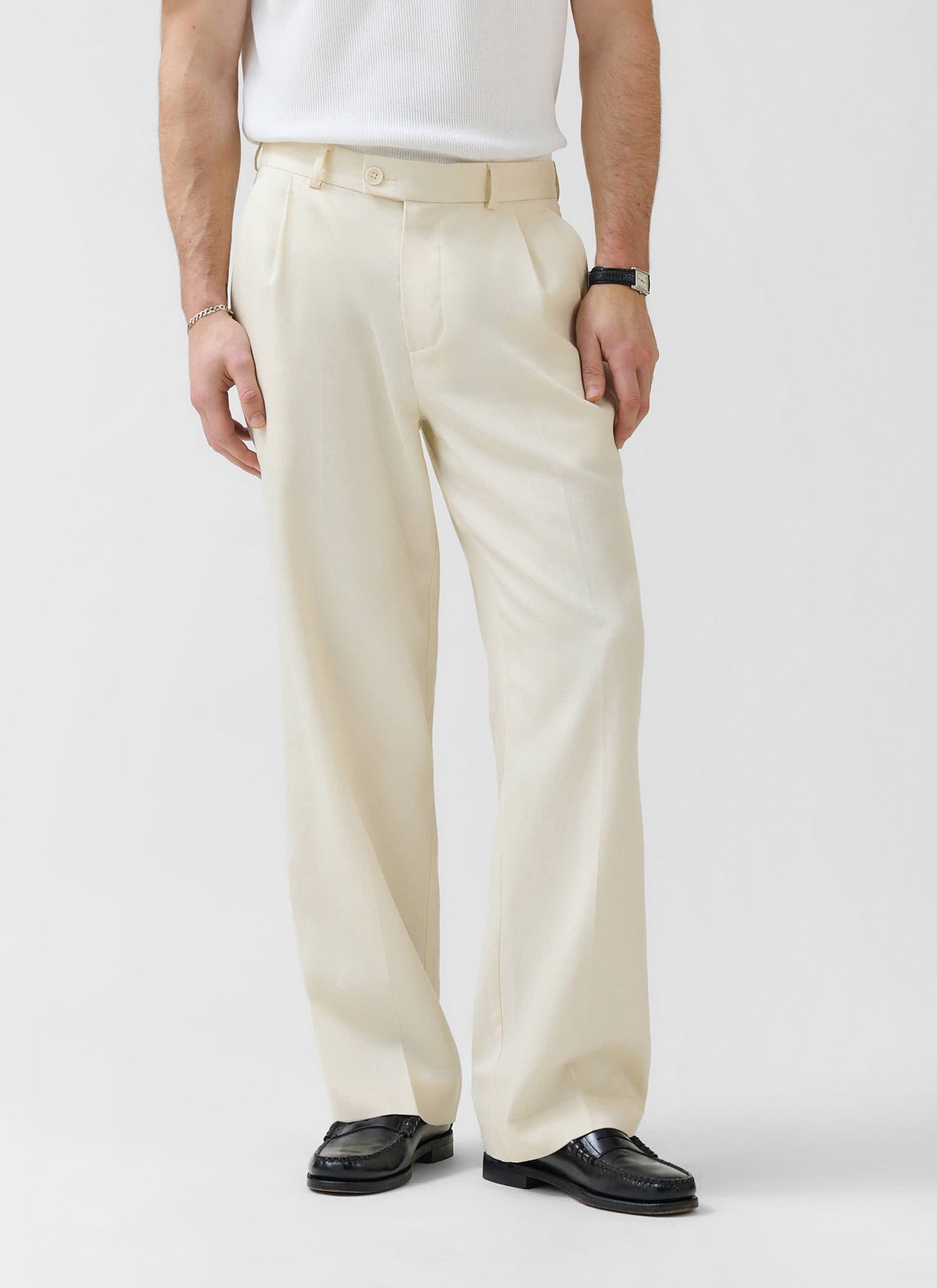 A model wears the Percival Pleated Tailored Trousers in cream with a white ribbed top and black loafers.