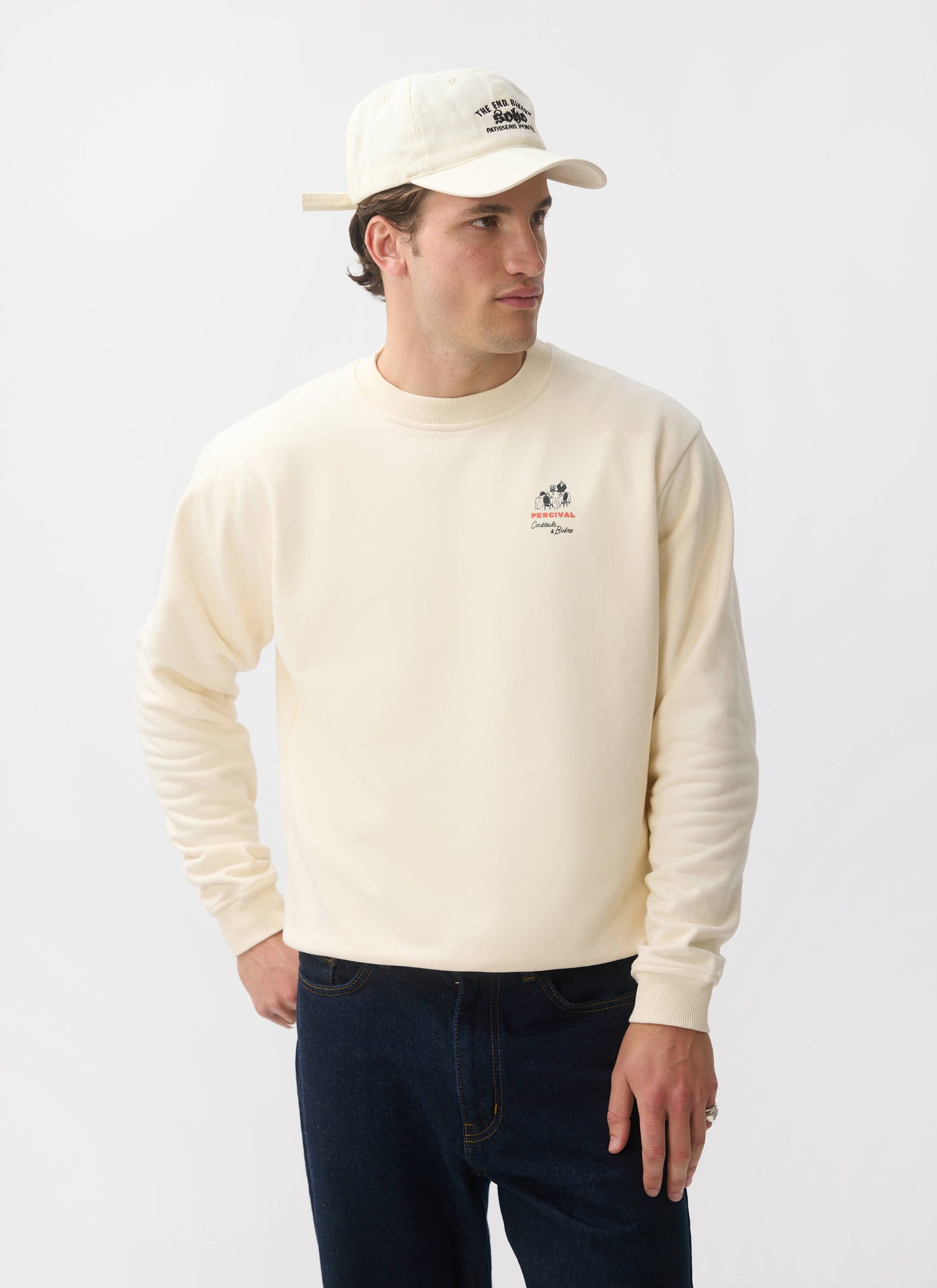 A model wears the Percival Bistro Printed Sweatshirt in ecru, styled with dark jeans and a baseball cap.