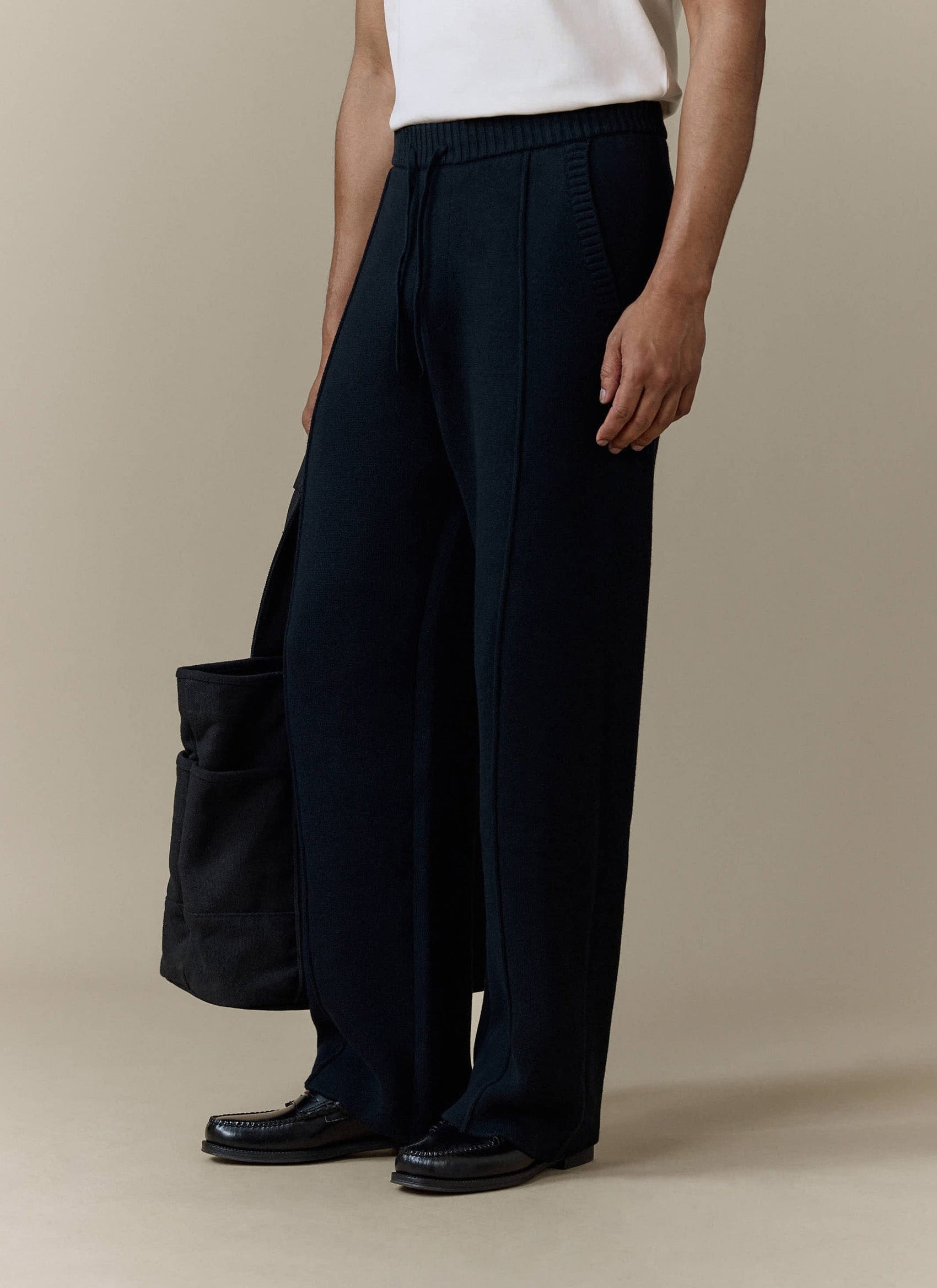 A model wears the black Chronicle Knitted Straight Leg Trackpants by Percival, styled with a tote bag and black loafers.