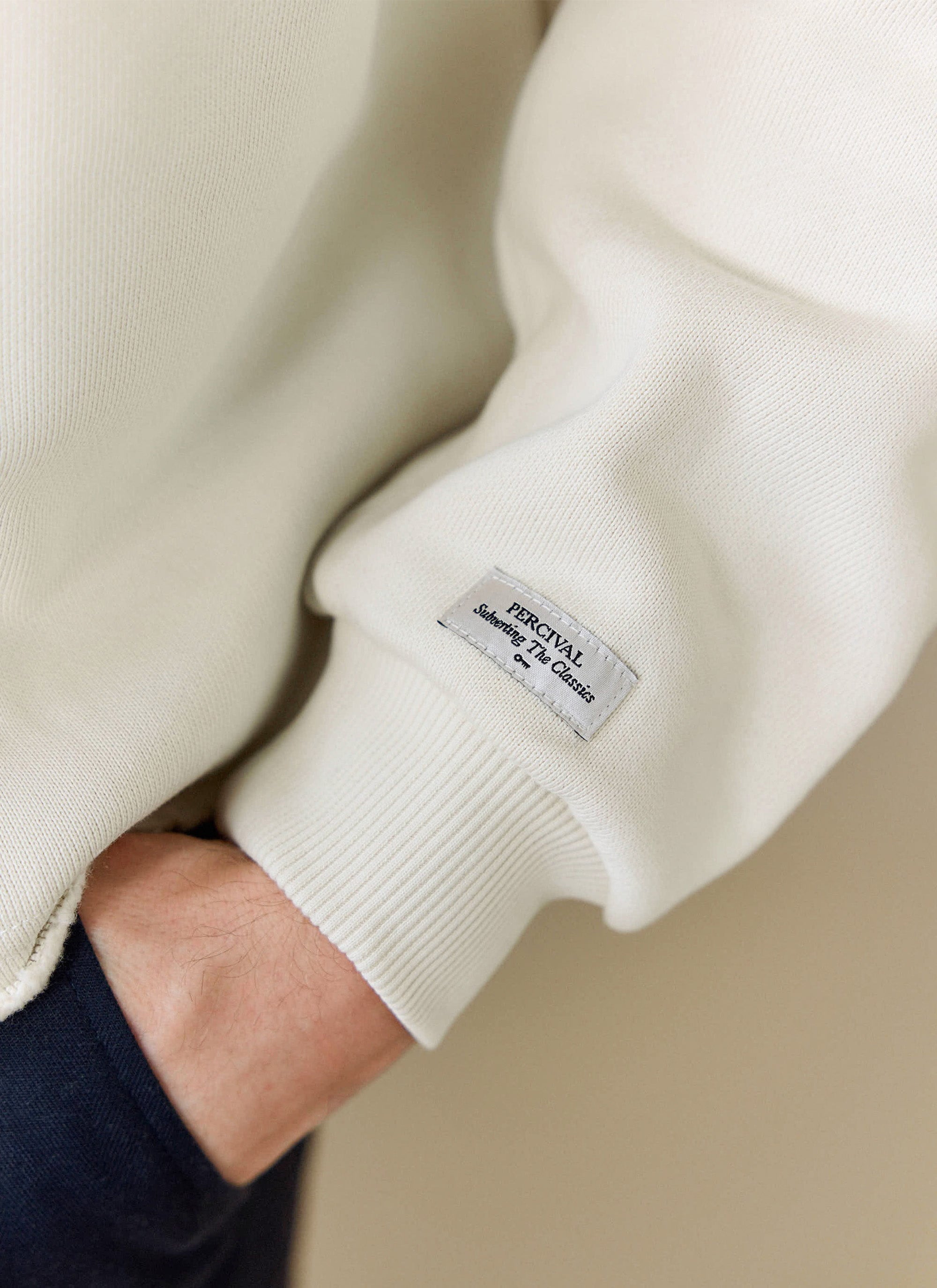 A close-up of the Percival Dudley Boxy Sweatshirt in ecru, showing the branded label on the sleeve above the ribbed cuff.