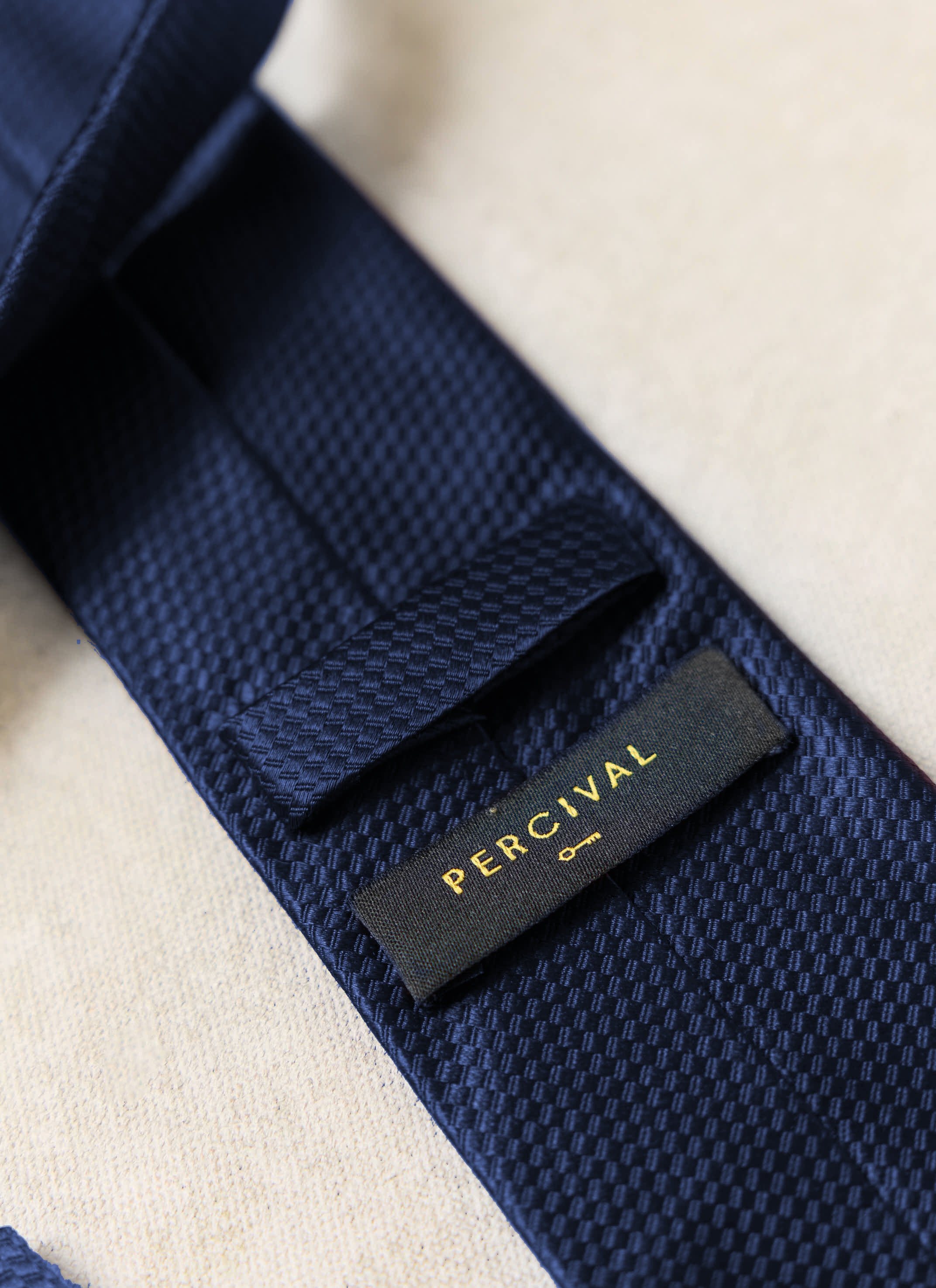 A close-up of the Percival Grenadine Tie in navy silk, showing the textured weave and black brand label.