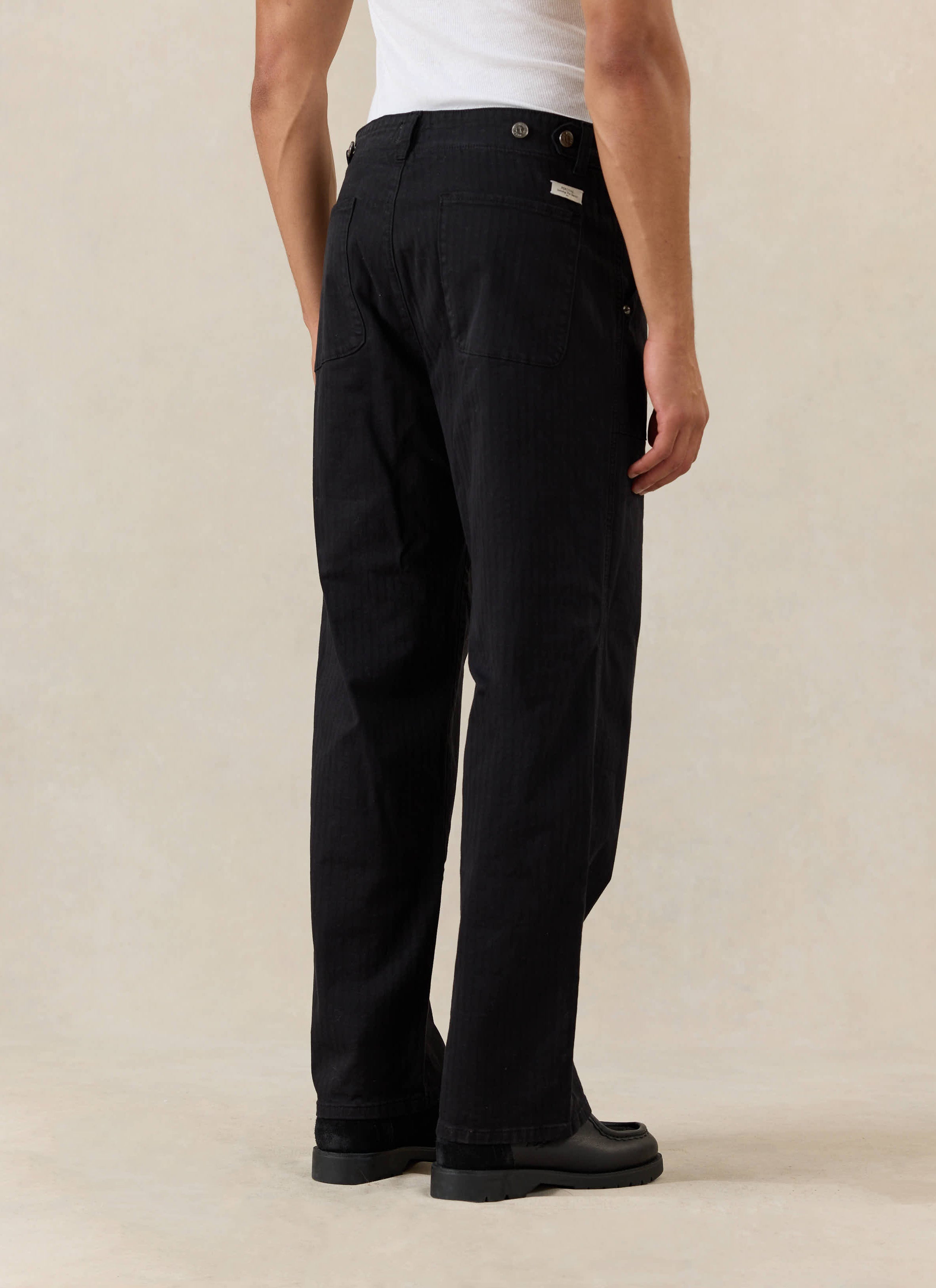 Rear view of the black cotton Lungo Carpenter Trousers from Percival, styled with a white vest and black loafers.