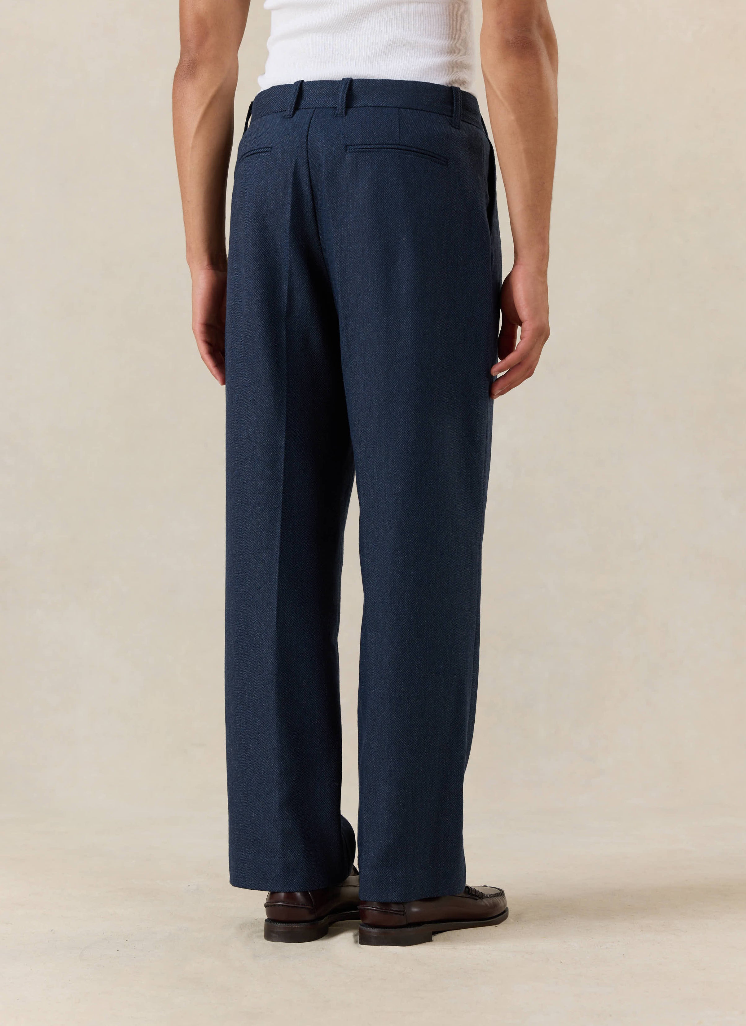 Percival Stanley Pleated Tailored Trousers in navy herringbone wool, shown from behind with a white vest and brown loafers.