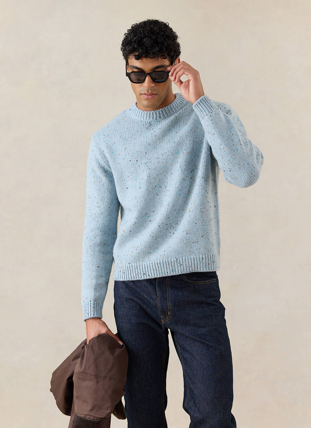 A model wears the blue Rhodes crew neck jumper from Percival, featuring a flecked texture and styled with dark jeans.