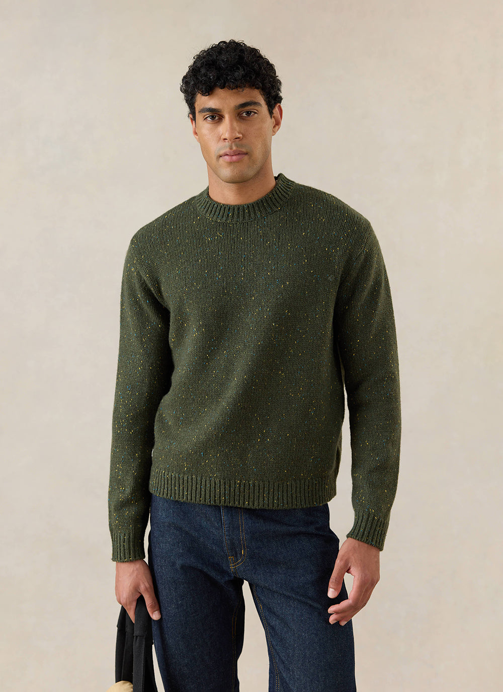 A model wears the forest green Percival Rhodes crew neck jumper, featuring a speckled texture, paired with dark denim jeans.
