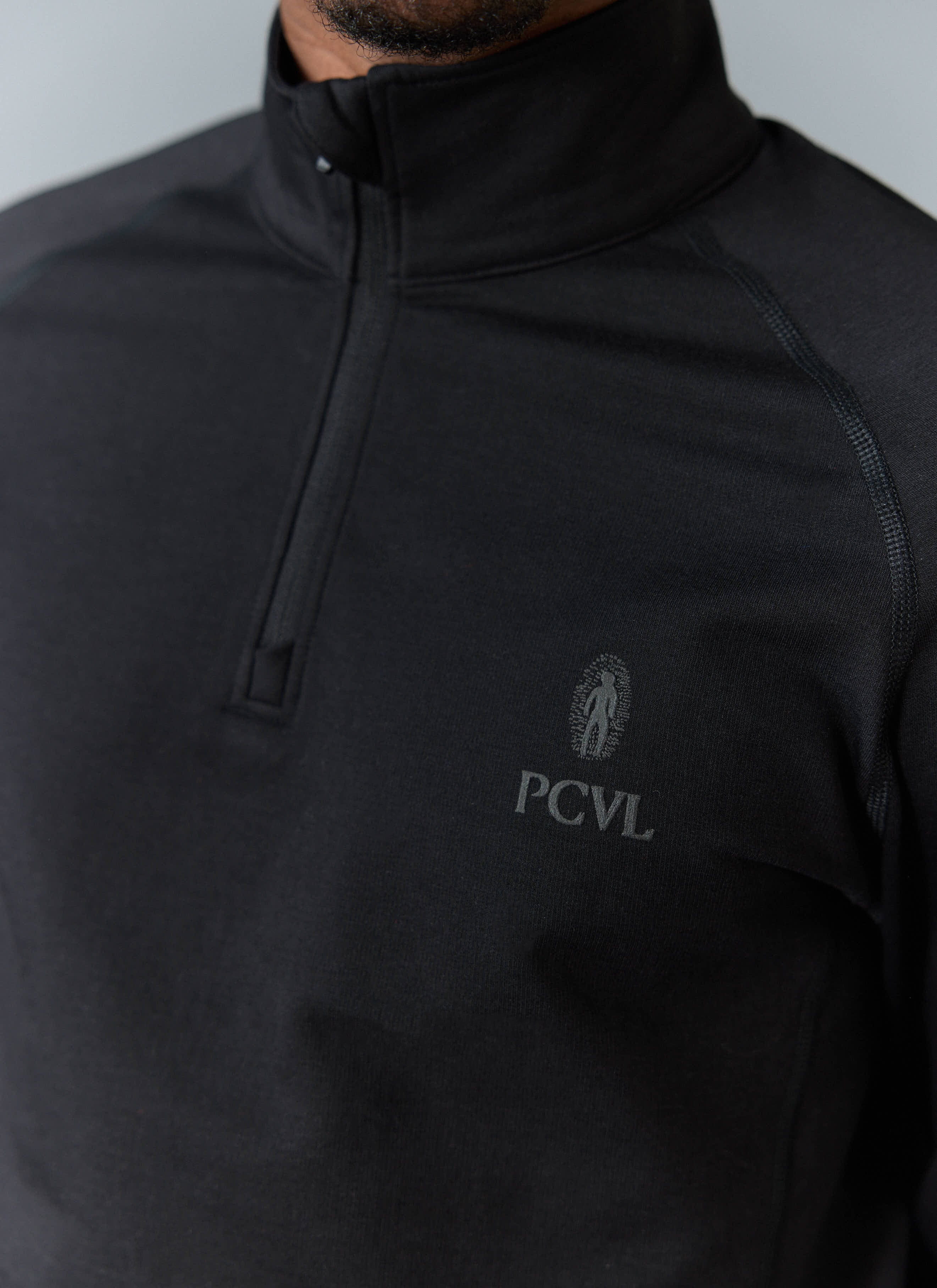 Close-up of a model wearing the black PCVL Base Layer Quarter Zip by Percival, showing the subtle reflective chest logo.