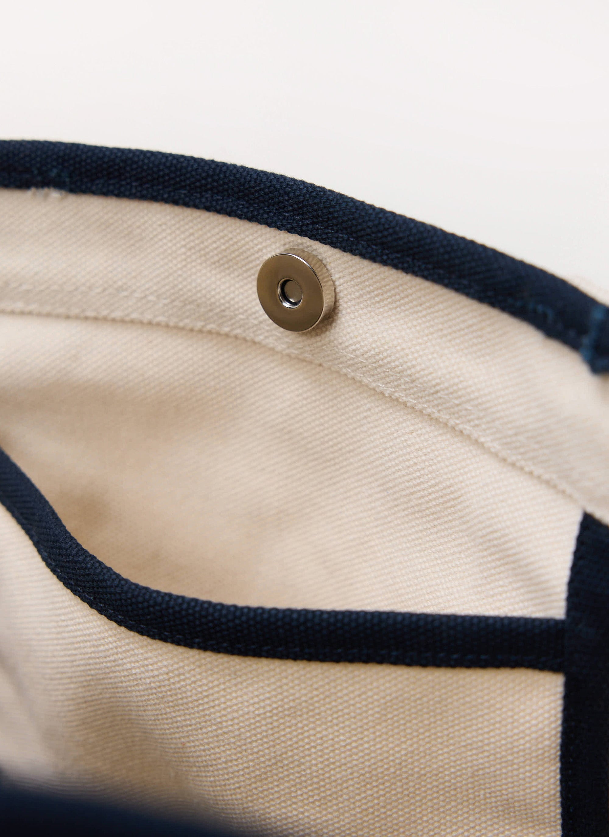 Close-up detail of the Percival ecru canvas tote bag, showing the navy trim, internal pocket and magnetic snap closure.