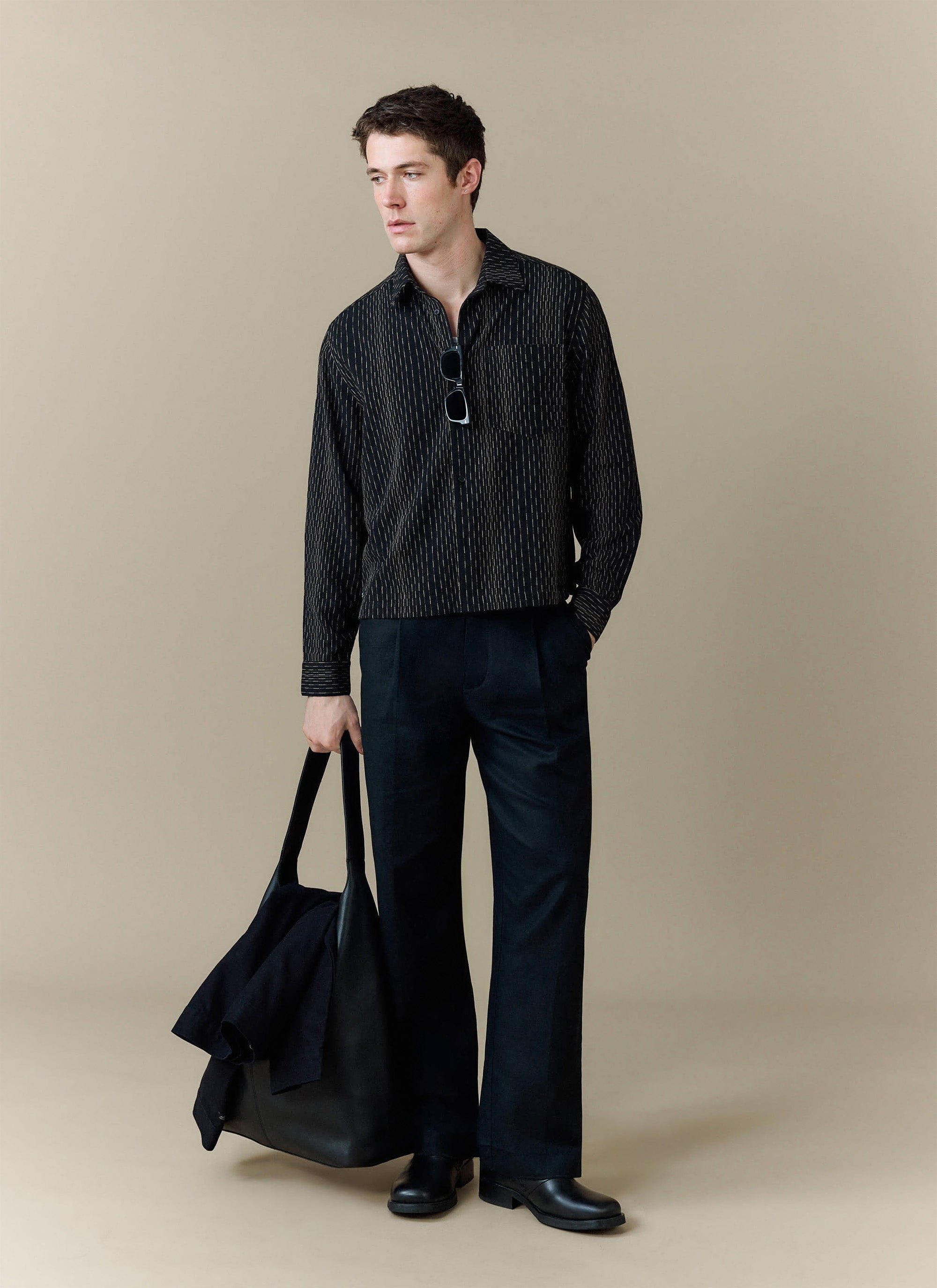 A model wears the black Leland Cuban Boxy Shirt from Percival, styled with black trousers and holding a large black tote bag.