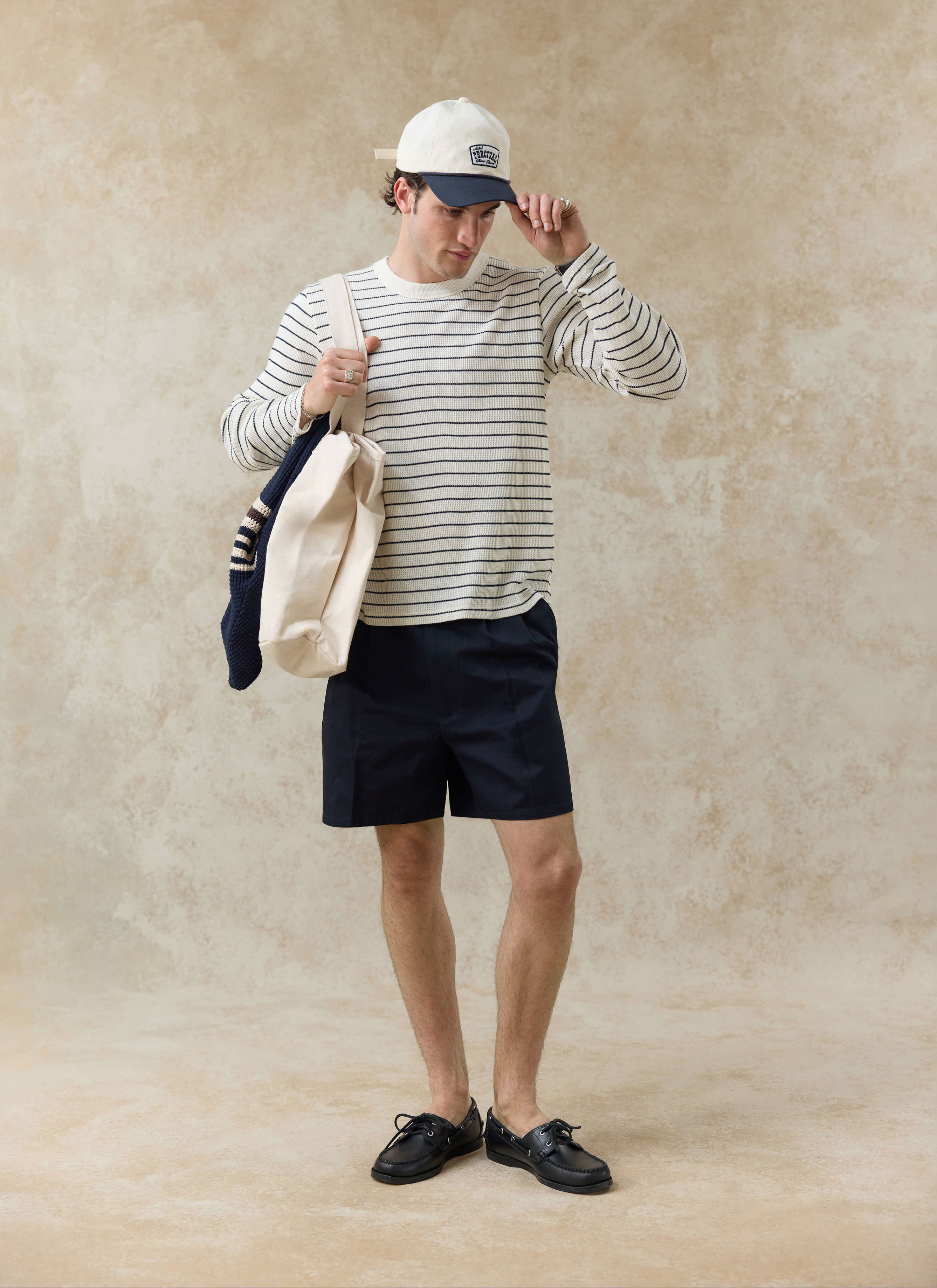 Model wears the Percival Ernie Waffle Long Sleeve T Shirt in cream and navy stripes with navy shorts and a tote bag.