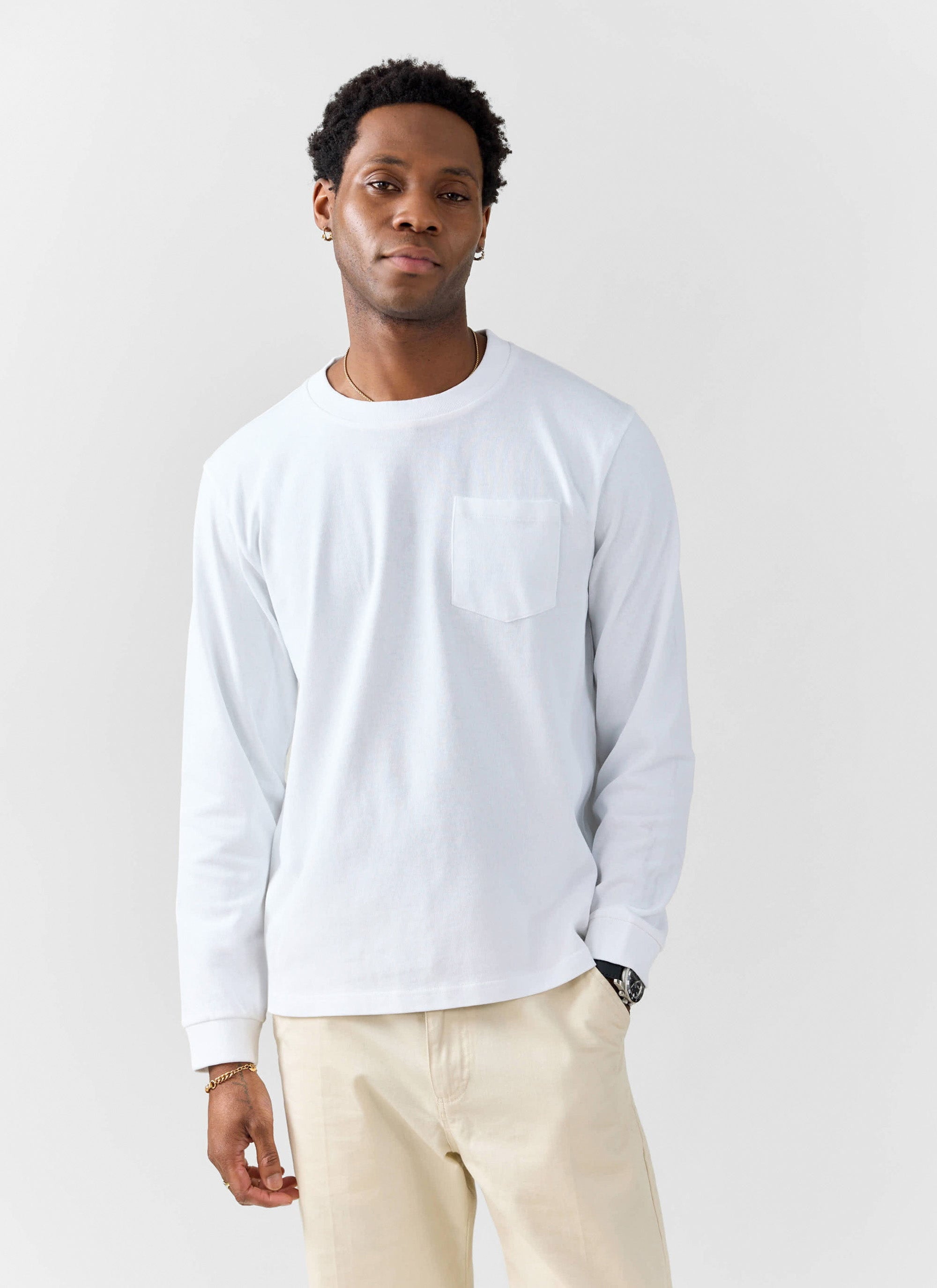 A model wears the Percival white Long Sleeve Pocket T-Shirt in organic cotton, paired with cream trousers.