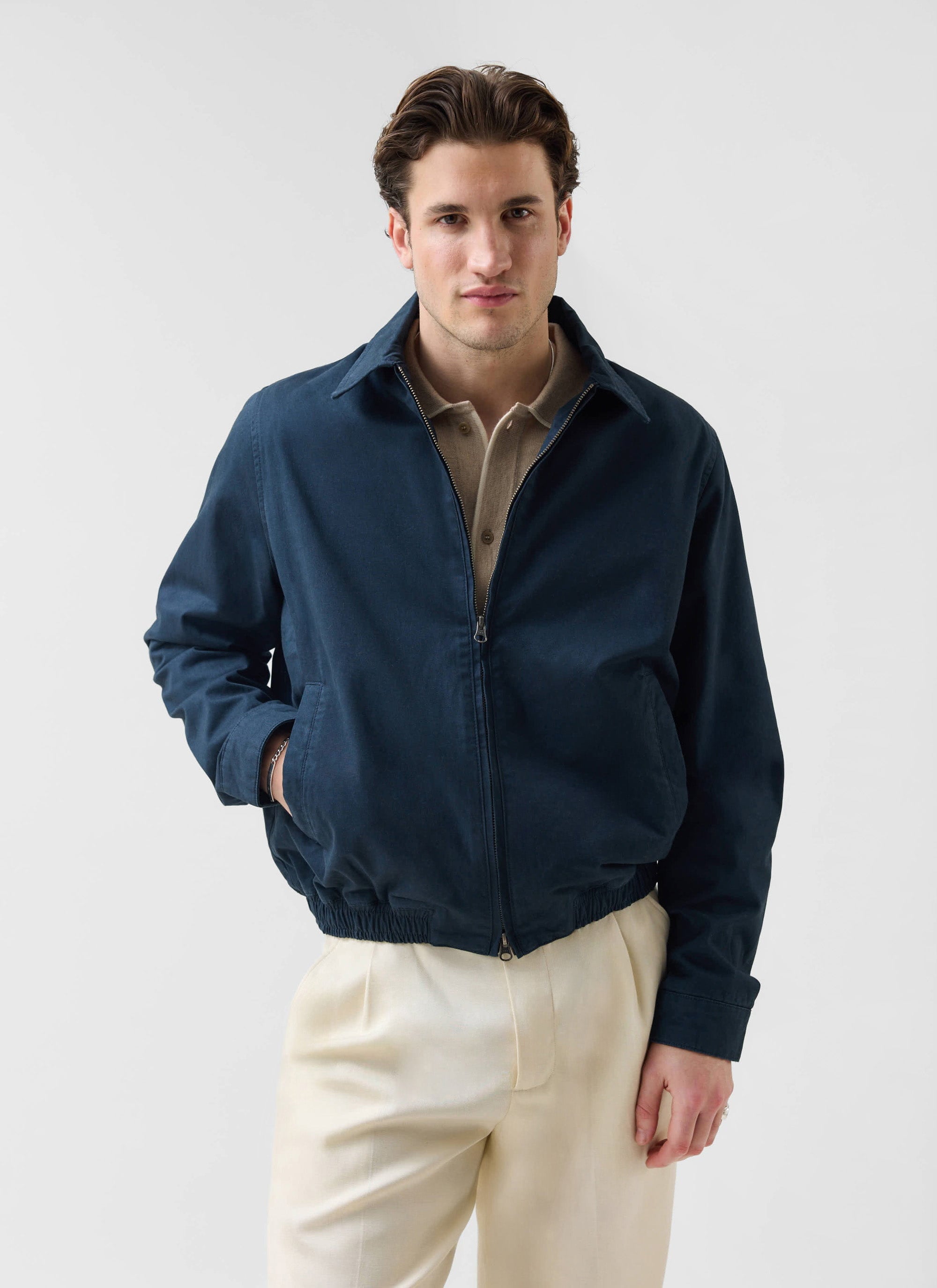 A model wears the Percival Leo Harrington Jacket in navy with a beige polo shirt and cream trousers on a white background.