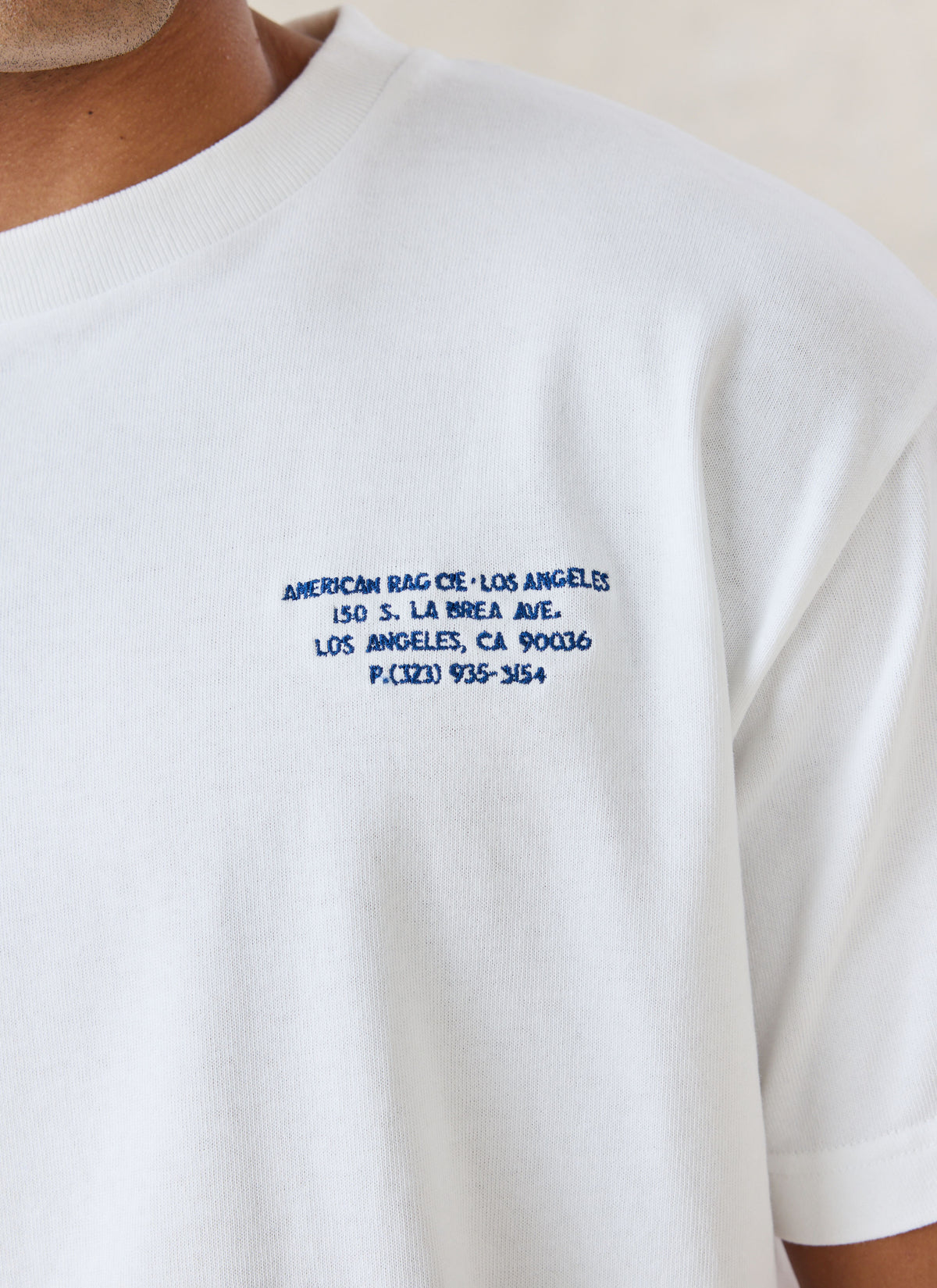 A close-up of the American Rag x Percival white oversized t-shirt showing the navy chest address lettering.