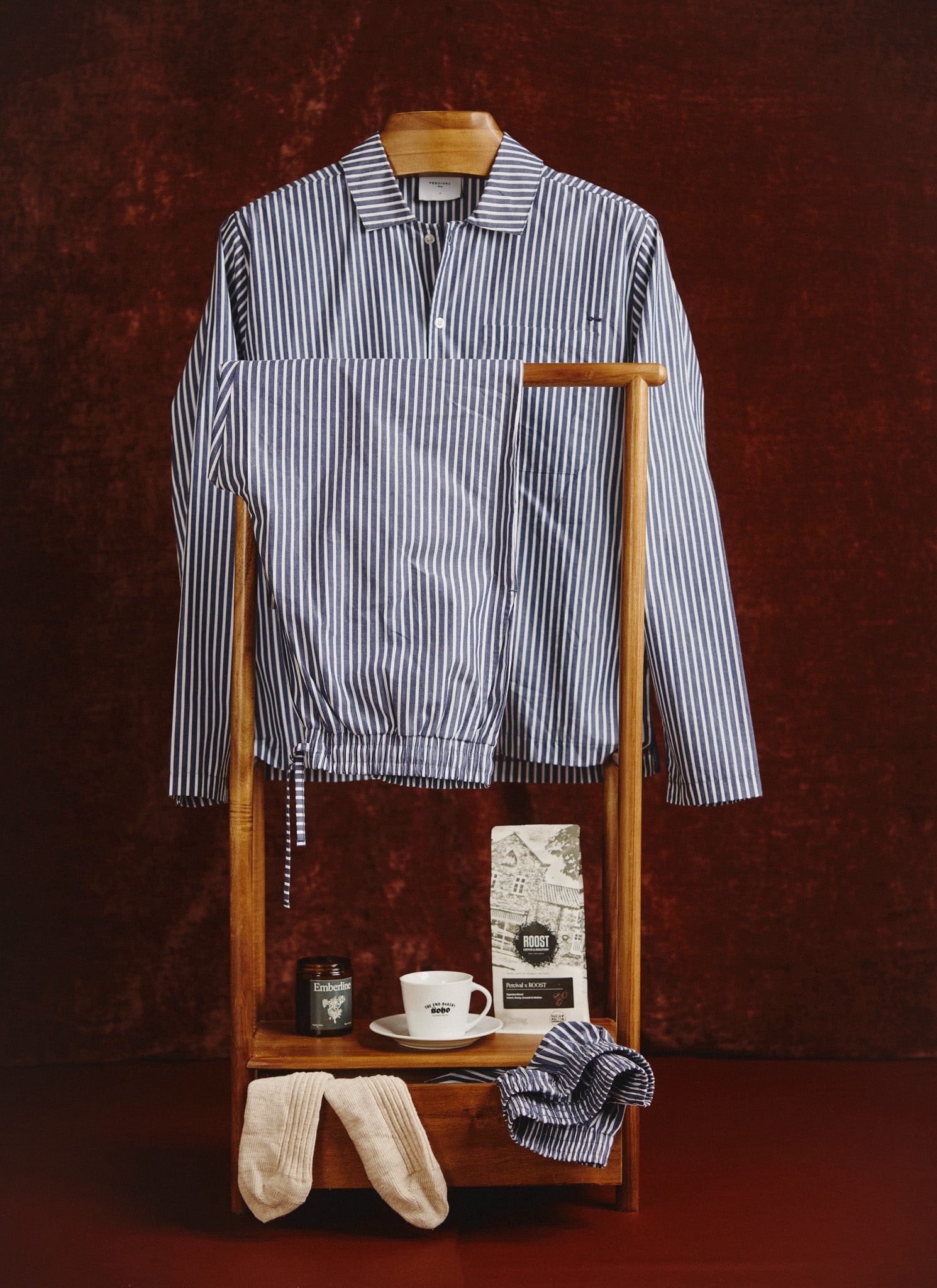 The Percival blue and white striped cotton pyjama set, displayed on a wooden valet stand against a rich brown wall.