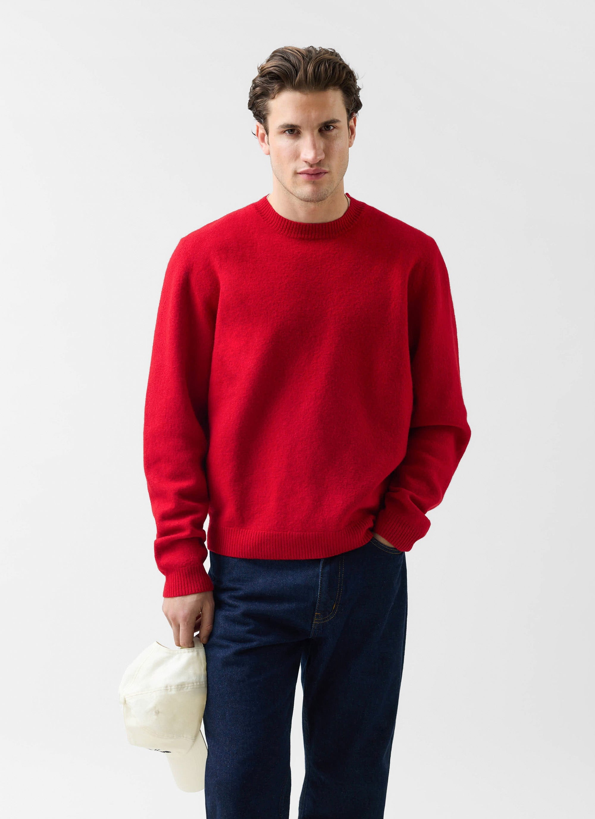 A man models the Percival Felted Regular Jumper in red merino wool, paired with dark blue jeans and holding a white cap.
