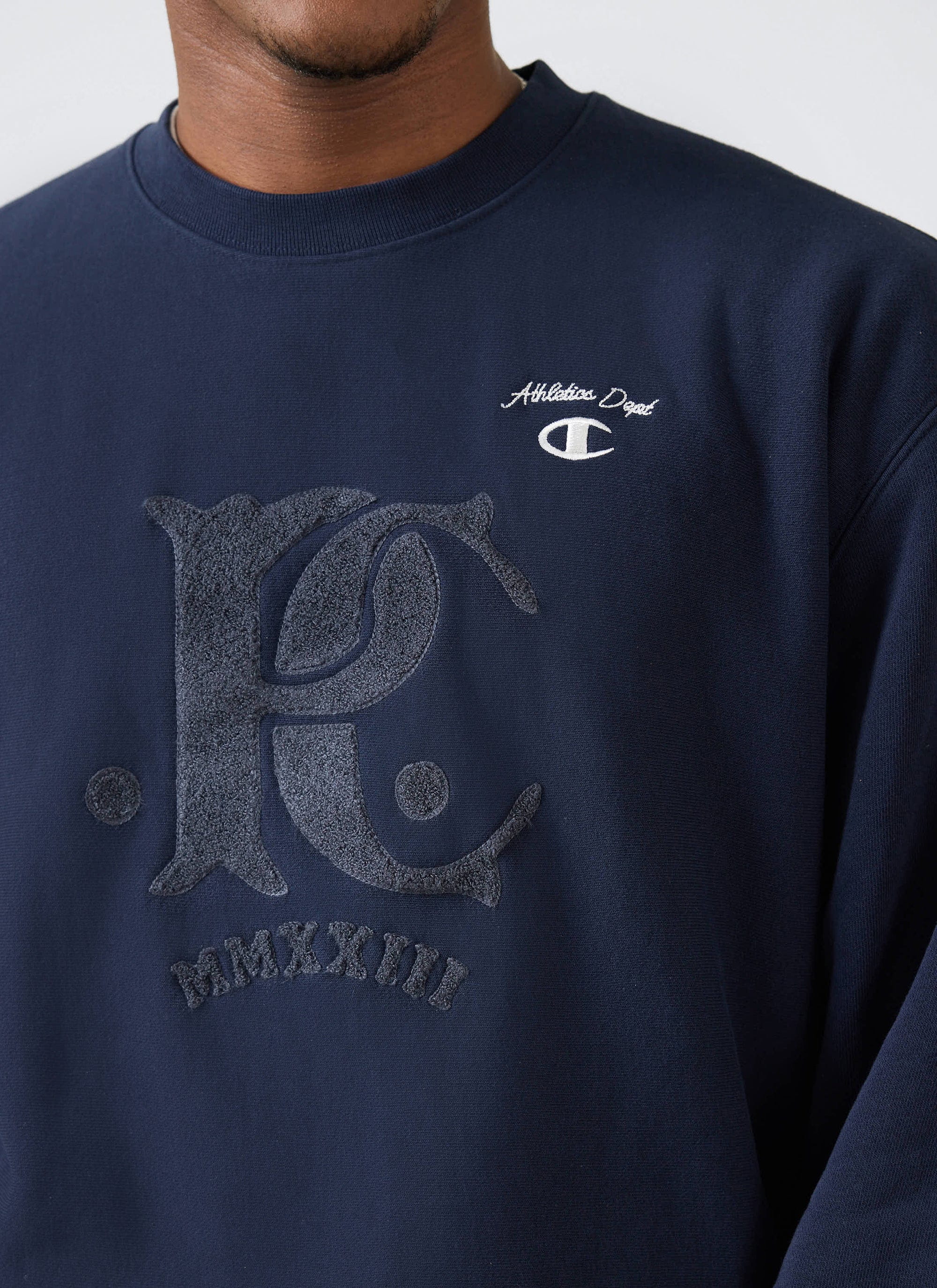 Close up of a model wearing the navy Champion x Percival sweatshirt, showing the tonal chenille patch detailing on the chest.