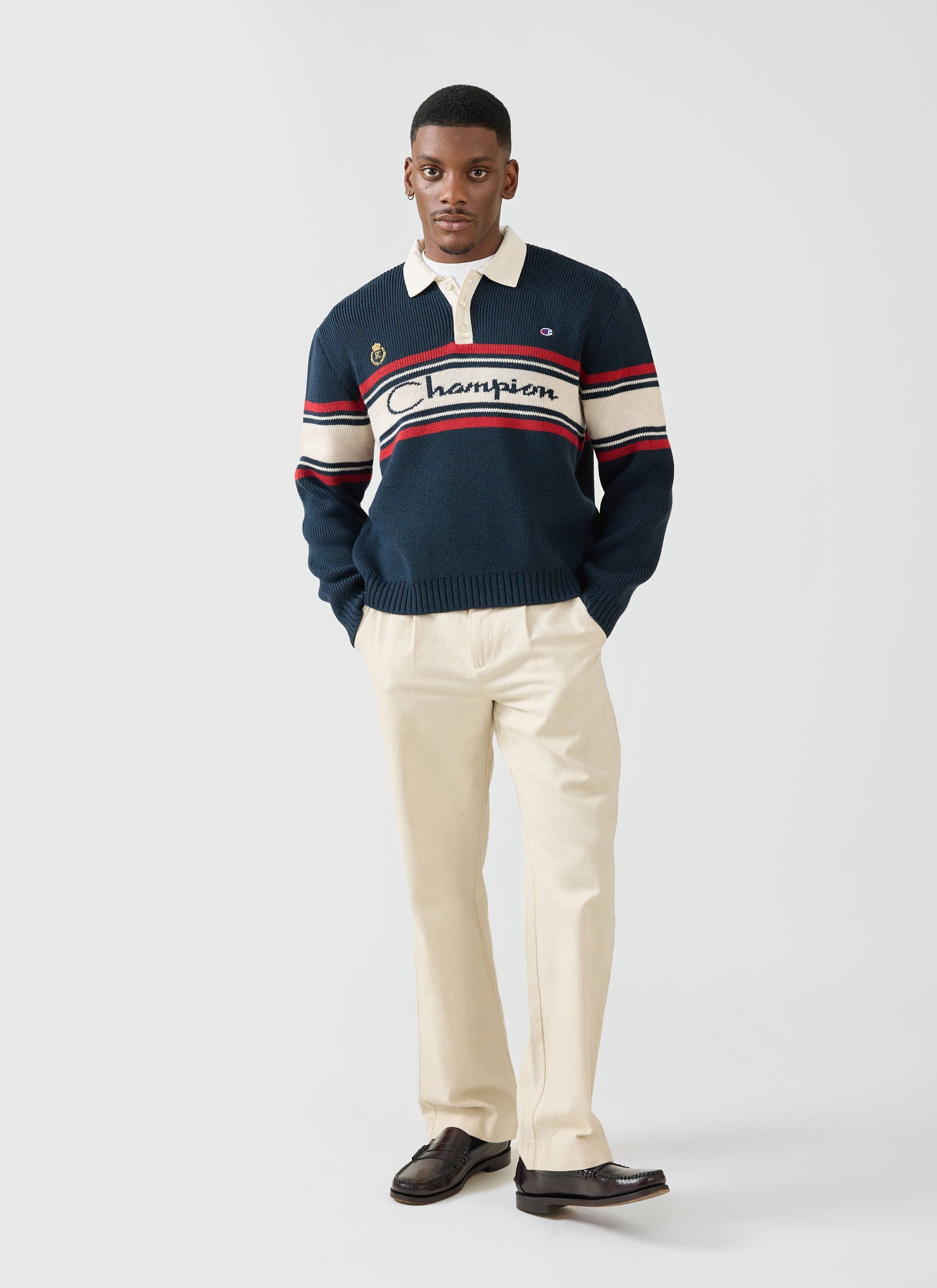 A model wears the Champion x Percival navy jacquard knitted rugby shirt with cream trousers against a white background.